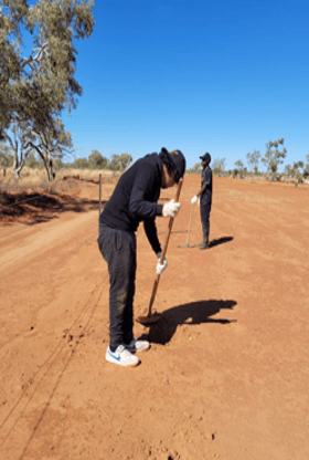 Two people on a dirt road surveying. One kneels, using a pole. Dry, reddish ground, clear blue sky.