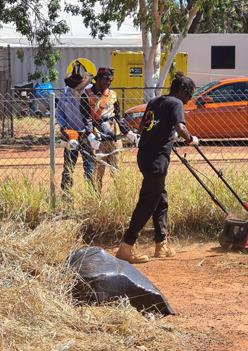 Three people, one mowing grass, two watching, in front of a fence and an orange car.