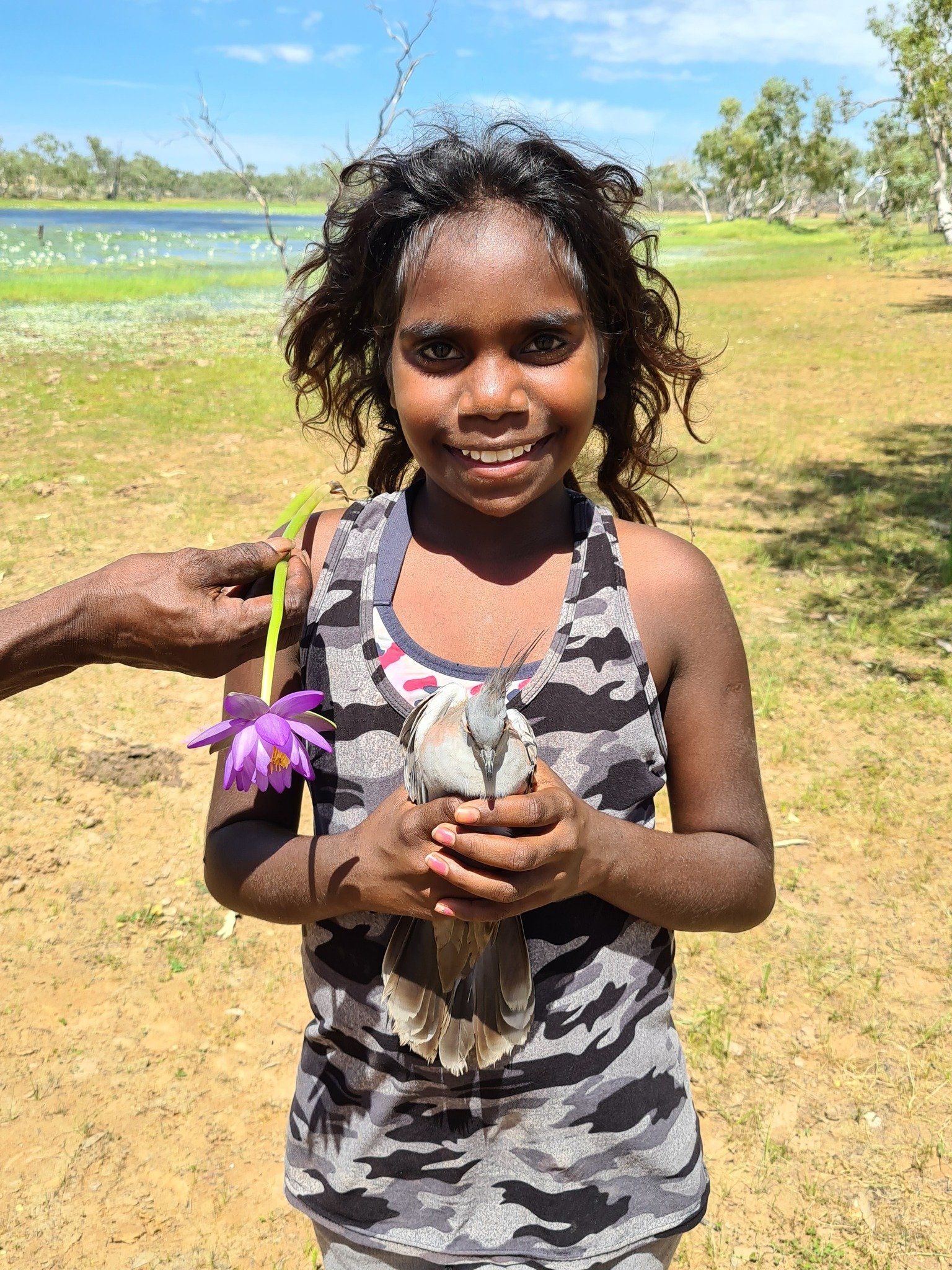 A young Indigenous girl smiles, holding a bird. She's outdoors near a lake, with a purple water lily offered to her.