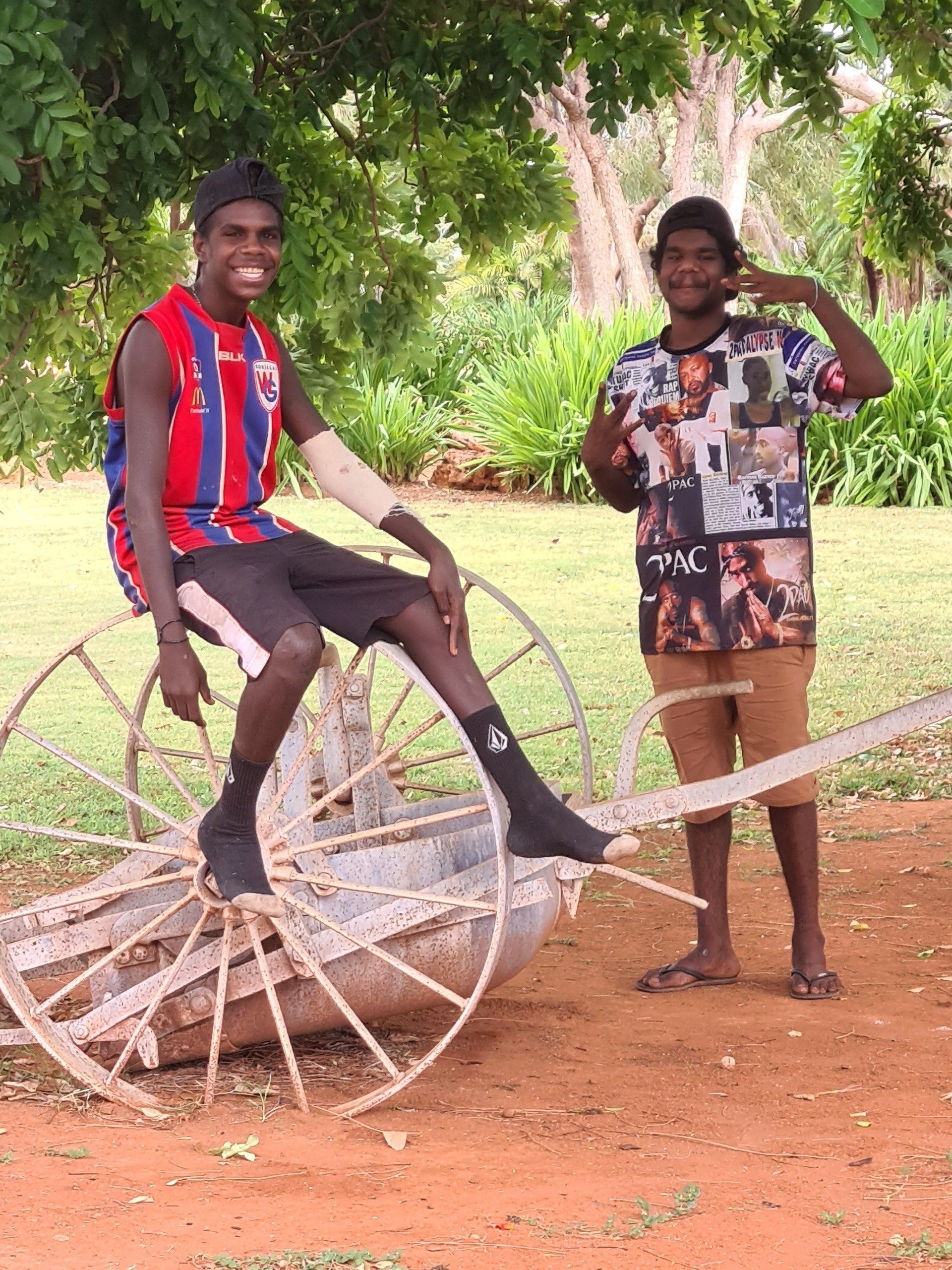 Two young Indigenous men in a park. One sits on a wagon wheel, the other makes a hand gesture.