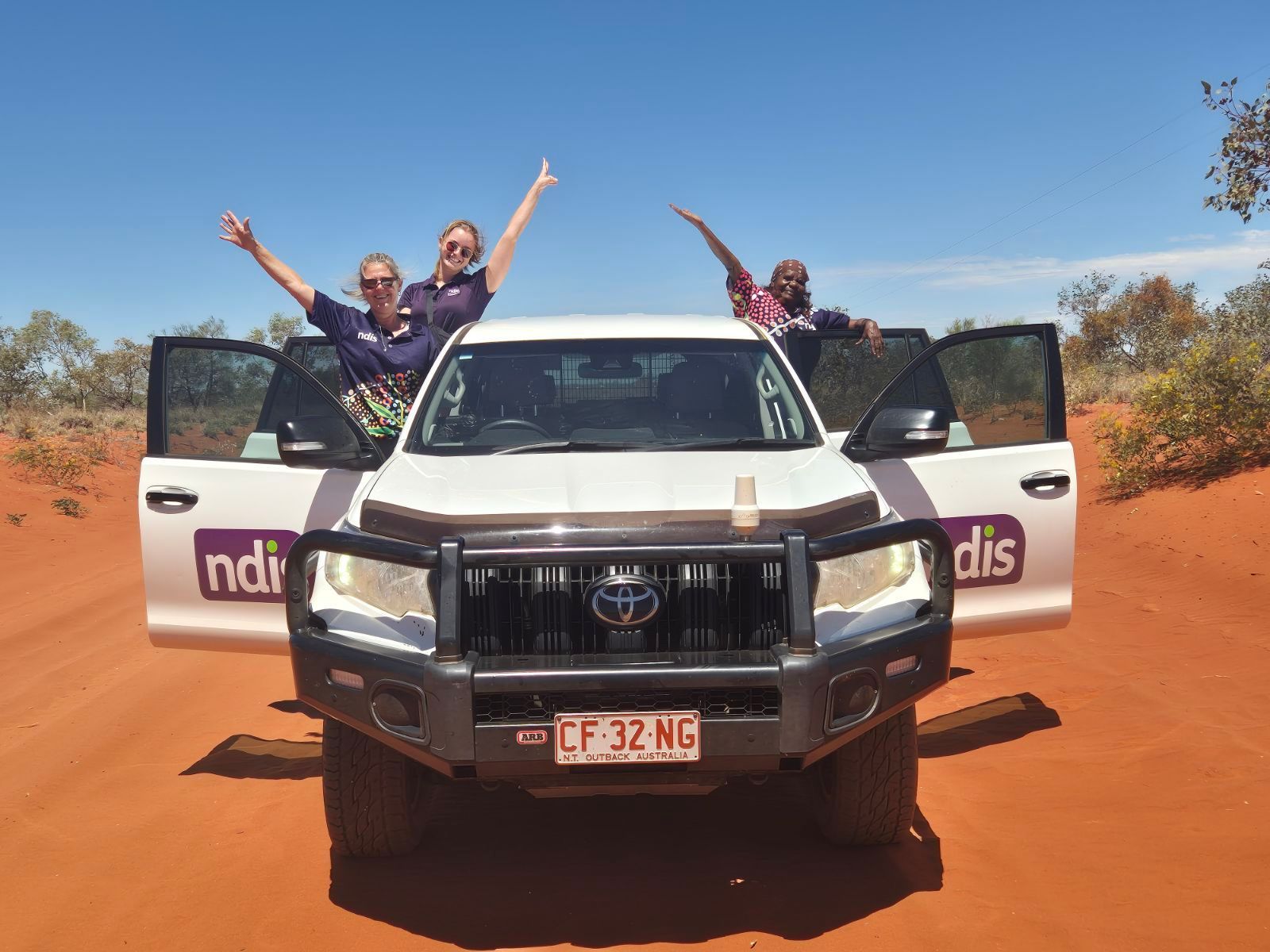Three people happily pose from a white SUV on a red dirt road under a bright blue sky. The vehicle's doors are open.