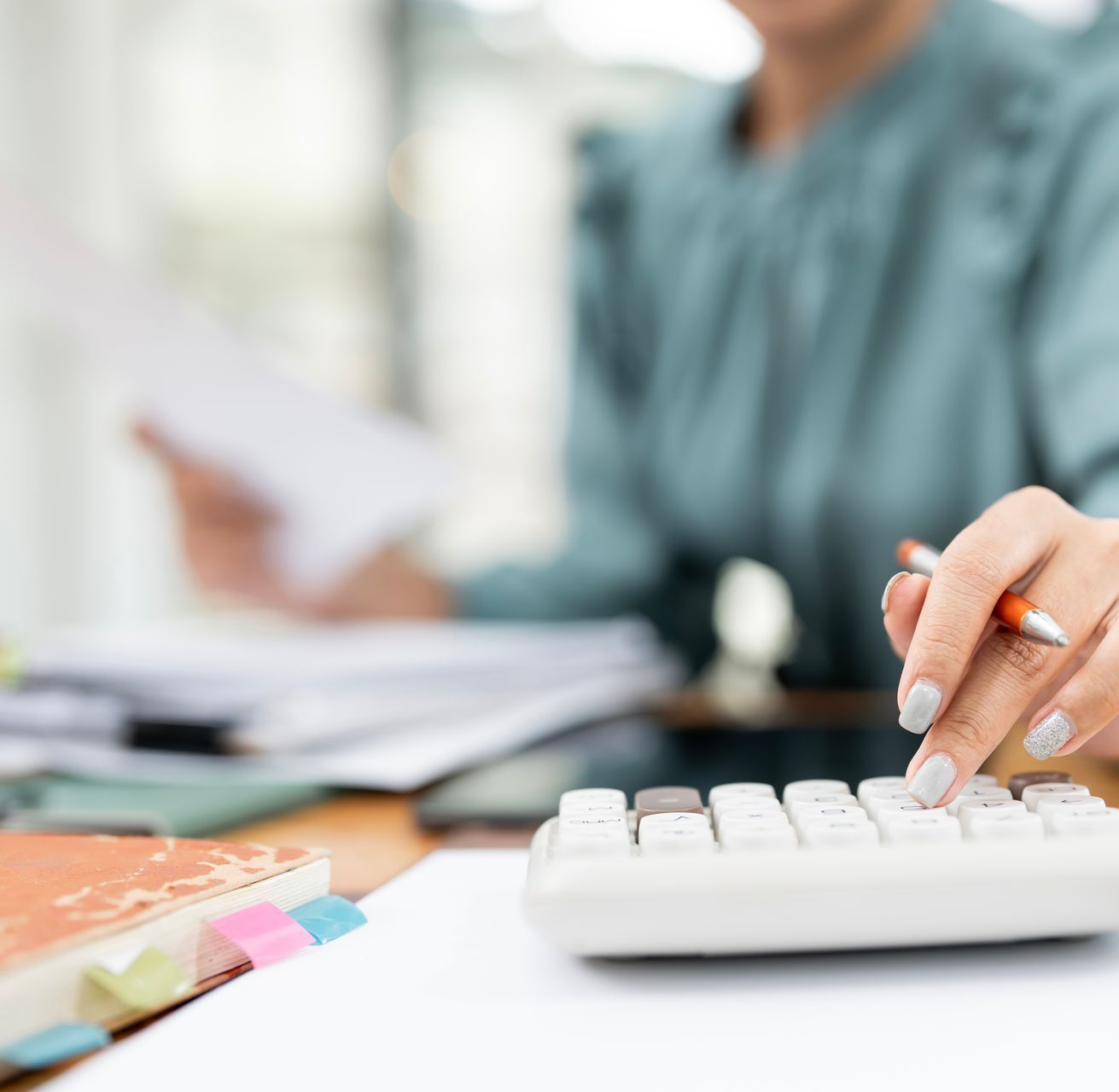 A woman is typing on a calculator while holding a pen.