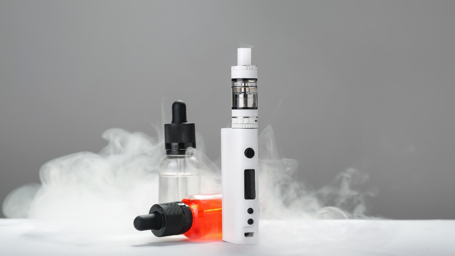A white vape mod with a clear tank, surrounded by two dropper bottles of e-liquid, emitting vapor in a smoky, gray background.