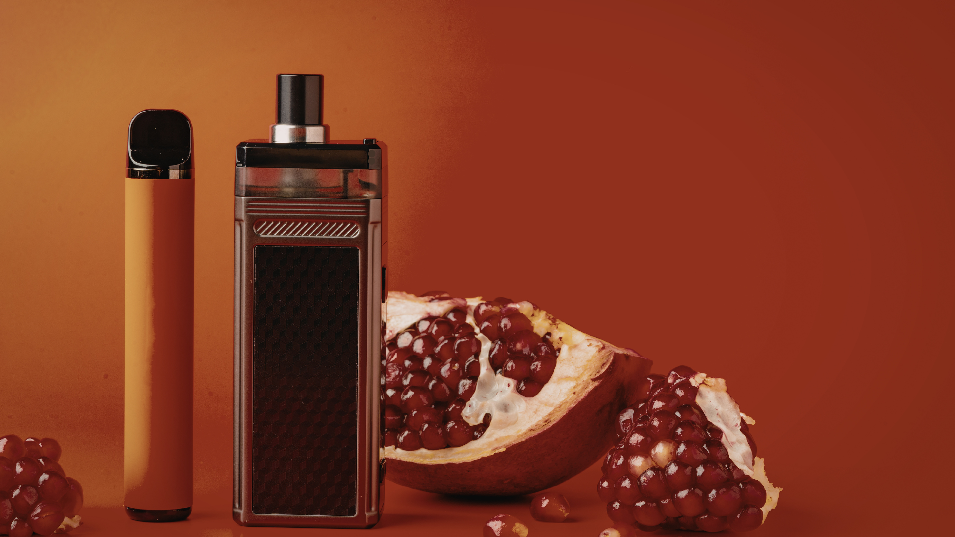 Two vape devices in front of a pomegranate with a rich orange background, highlighting a sleek, beginner-friendly pod system and a disposable vape.