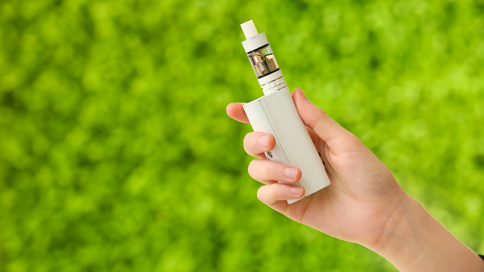 A hand holding a white vape mod with a clear tank against a vibrant green blurred background.