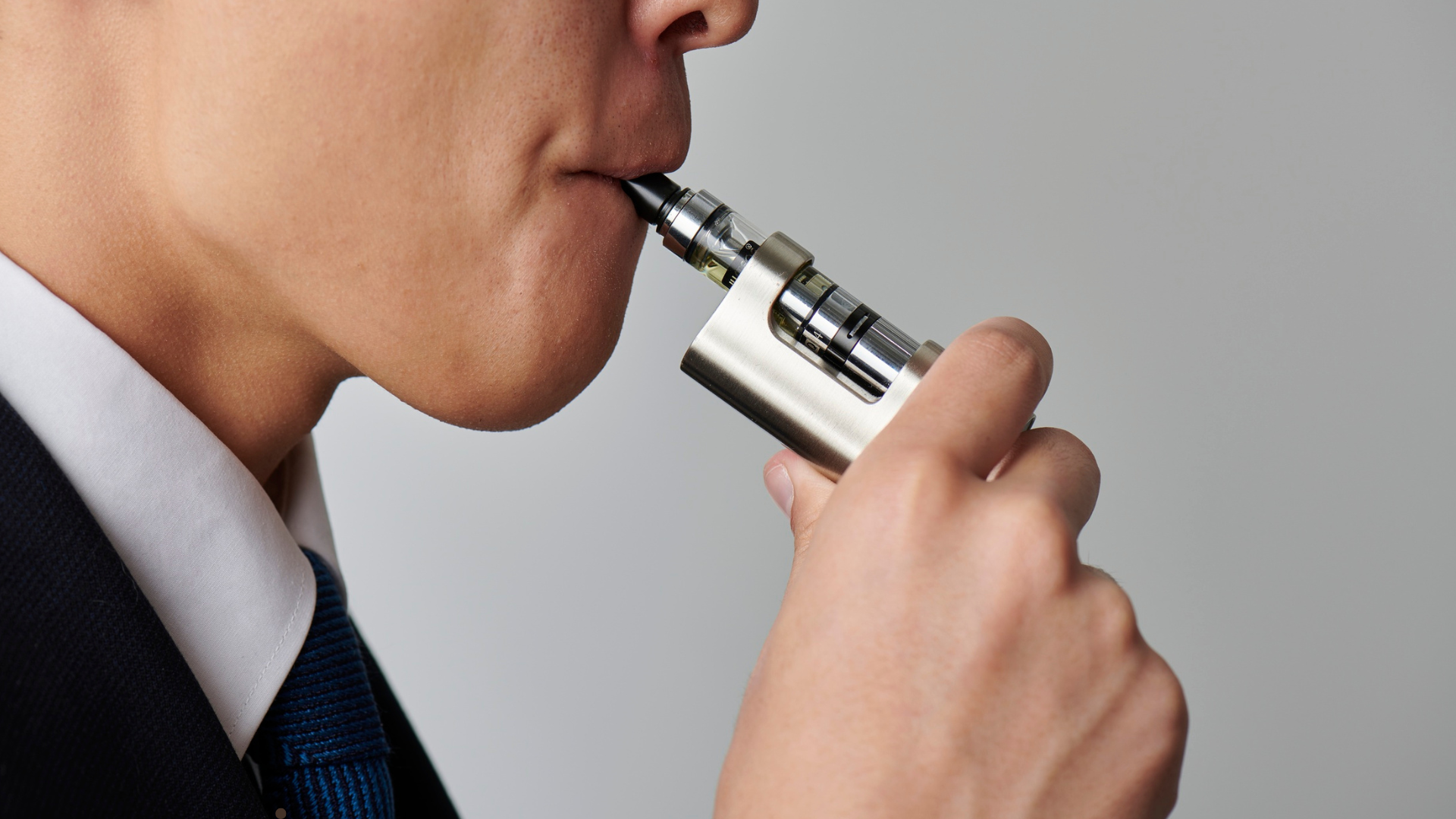 A person in a suit is holding a sleek, metallic vape device and inhaling from it, with a close-up vi