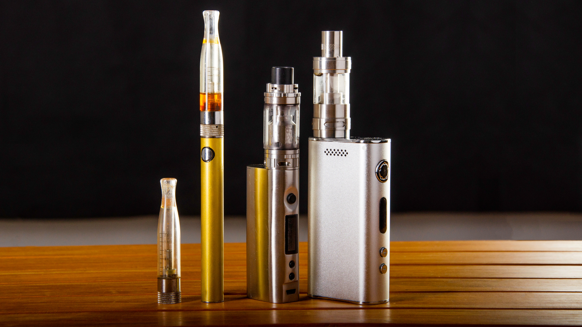 Various vape mods, including a gold pen-style vape, silver box mods, and clear tanks on a wooden table with a black background.
