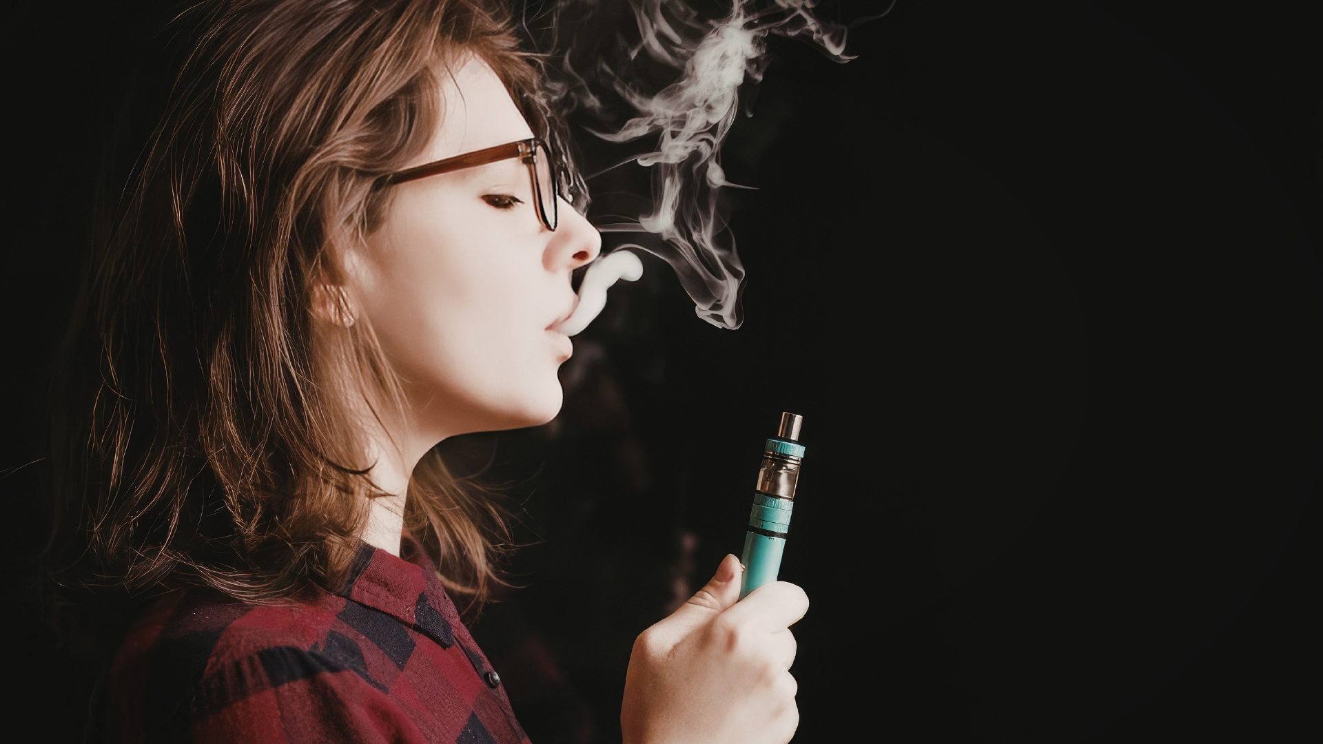 A woman exhaling vapor while using a teal-colored vape device, with a dark background highlighting the vapor cloud.