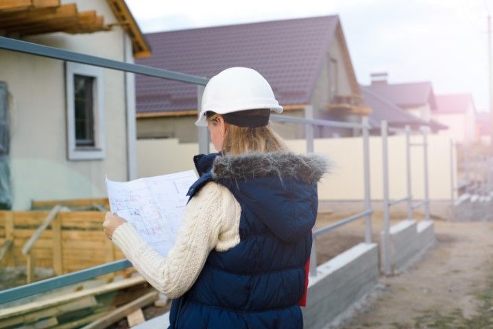 Woman in hard hat and vest reviews blueprints at a construction site, houses in the background.