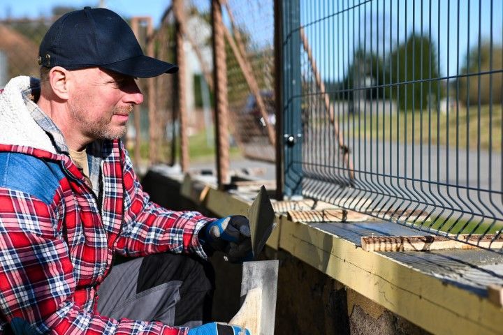 Man installing a wire fence, squatting outdoors. He wears a plaid jacket and baseball cap, looking at the work.