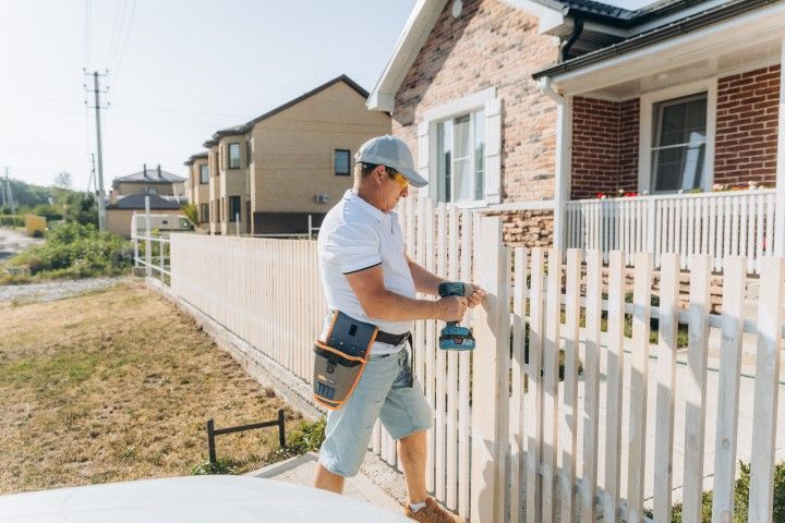 Man in hat installing a white fence with a power drill outside a house.