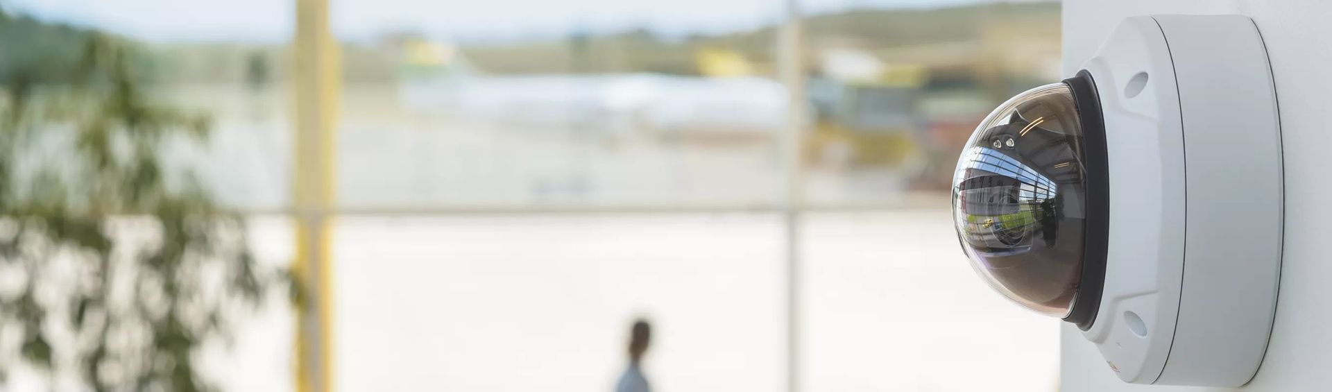Security camera mounted on a white wall, overlooking a blurred interior with a person in the distance.