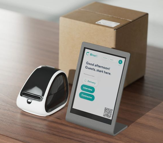 White and black robot-like device, tablet, and cardboard box on a wooden surface. Tablet displays a welcome message.