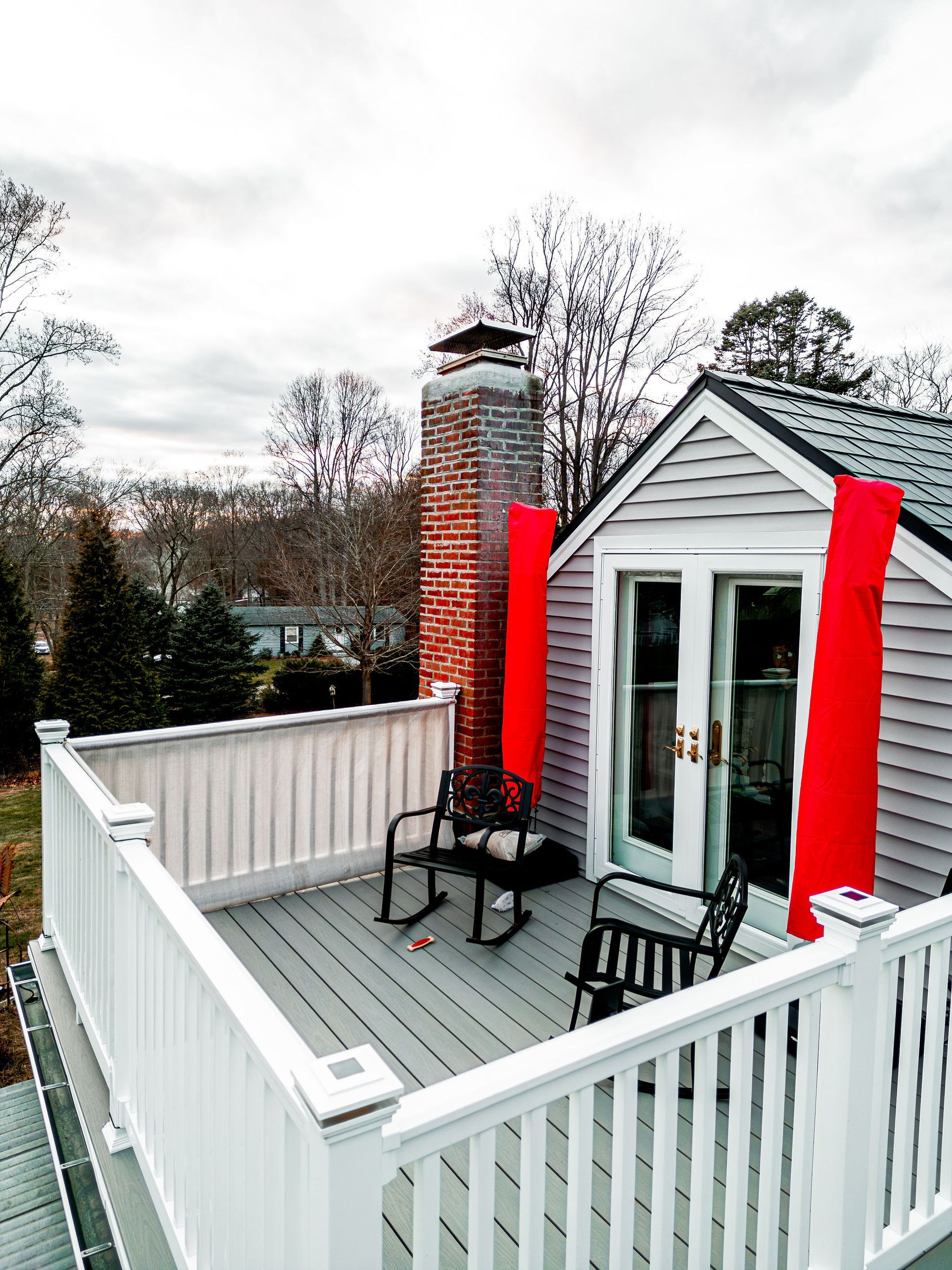 Rooftop deck with rocking chairs, brick chimney, gray siding, and red fabric against a cloudy sky.