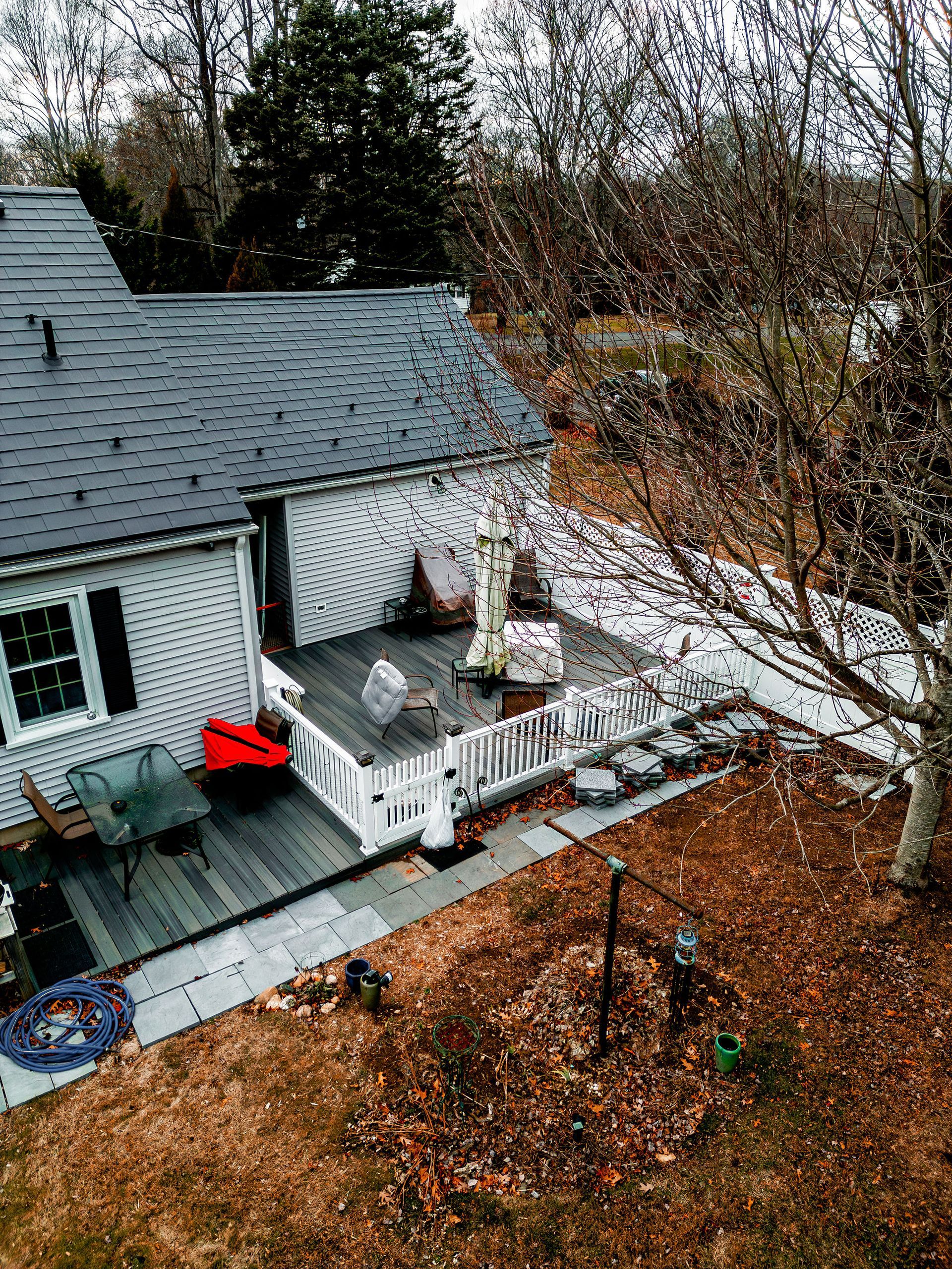 House with deck and yard. Gray roof, white siding. Deck furniture visible. Brown yard with bare tree.