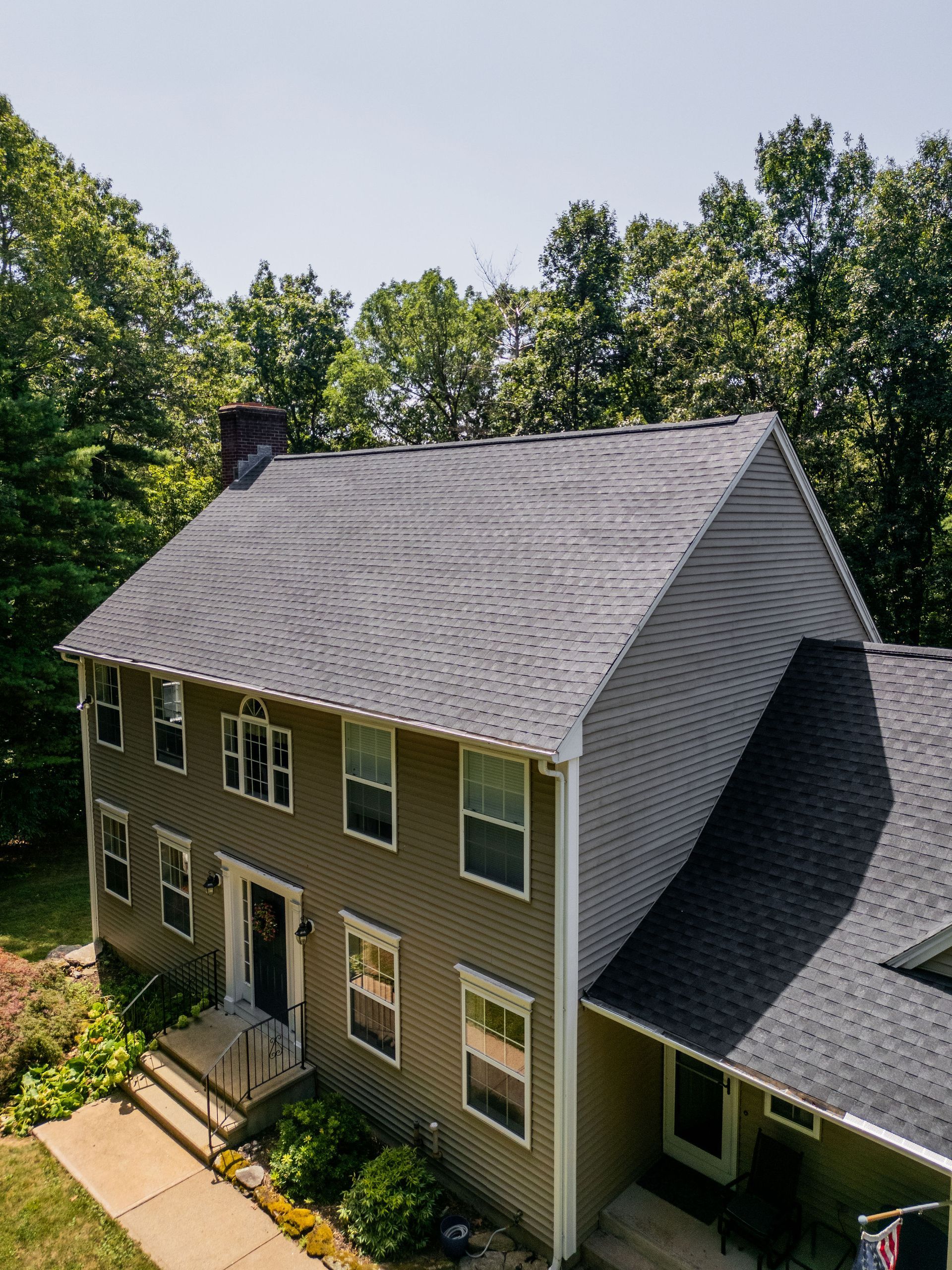 Two-story house with dark roof and light siding, surrounded by green trees under a clear sky.