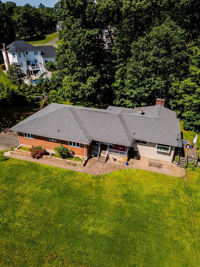 Aerial view of a single-story house with a gray roof, red brick facade, and a green lawn surrounded by trees.