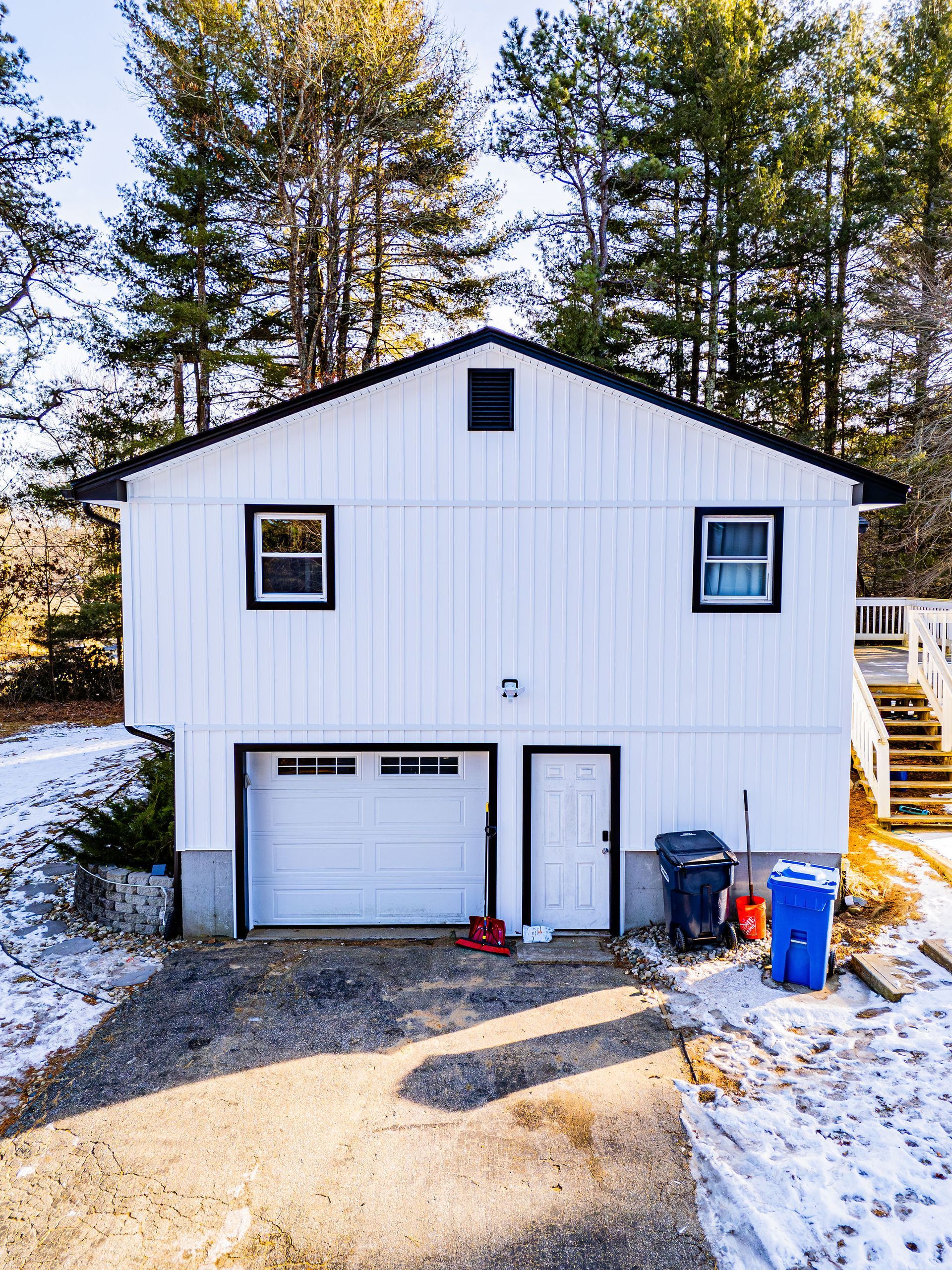 White two-story house with garage, door, and windows, driveway with snow. Trees in the background.