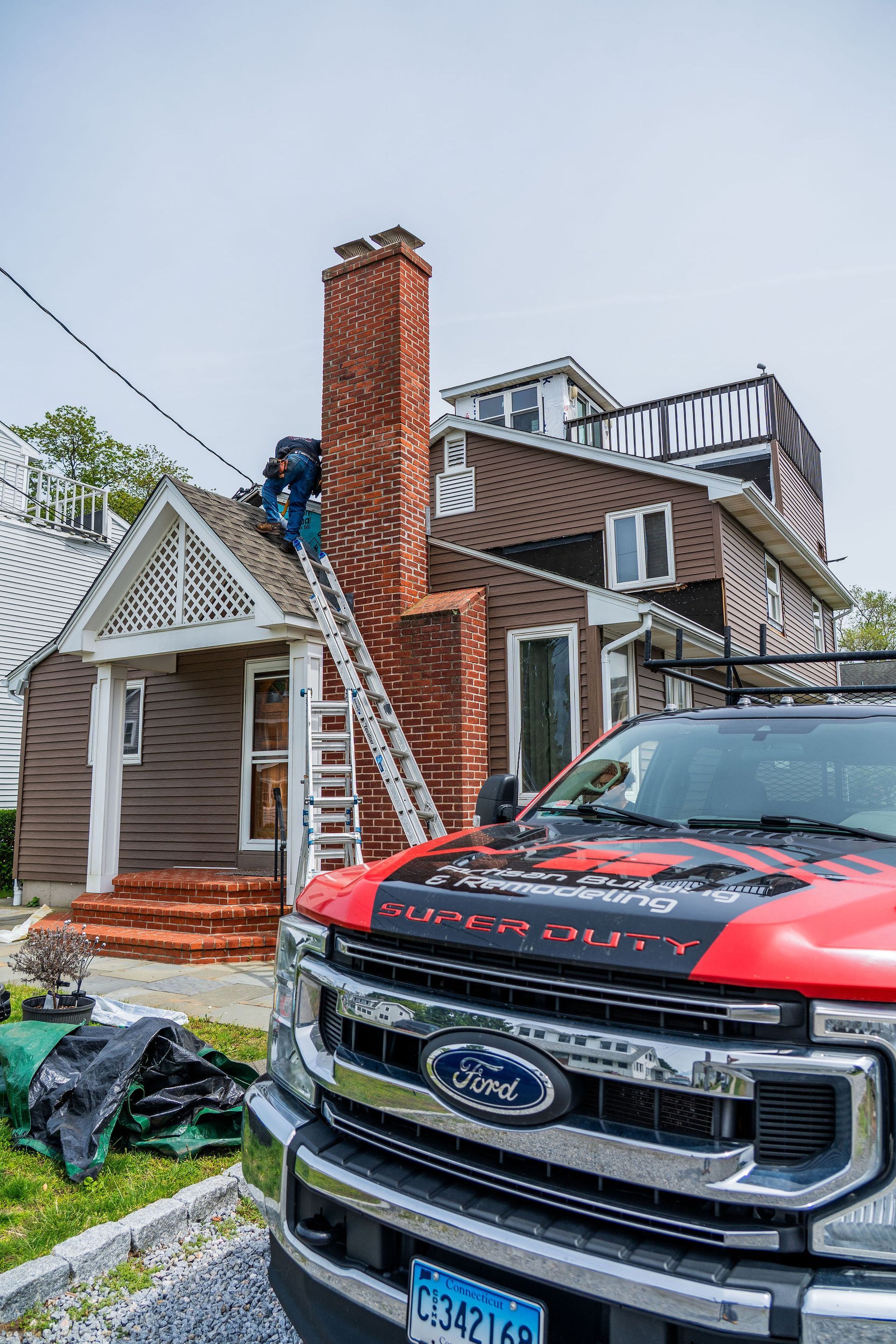 A worker on a ladder repairs a brick chimney on a brown house; a red Ford truck is in the foreground.