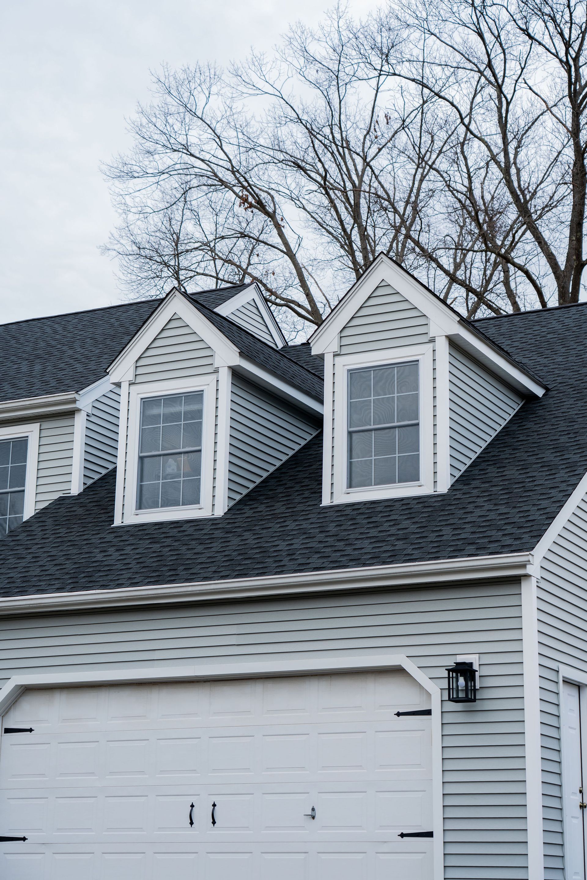 Gray house with white trim, three dormers, and a garage door. Bare tree branches in the background.