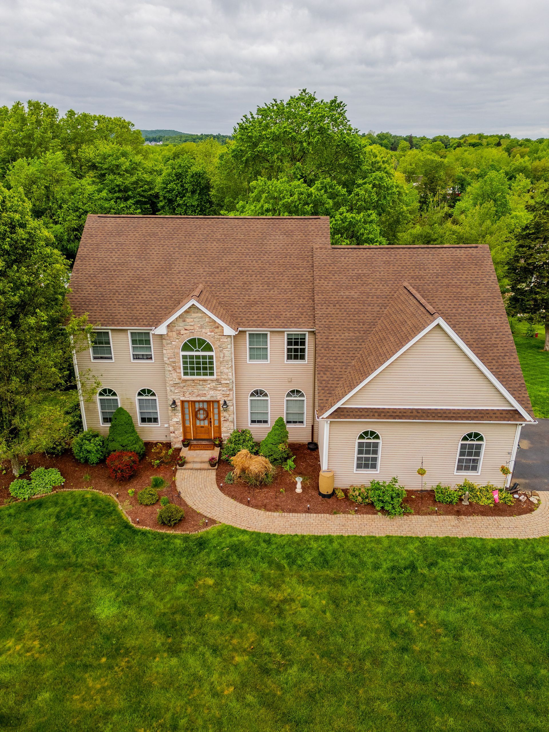 Two-story beige house with brown roof, stone entrance, and green lawn surrounded by trees under a cloudy sky.