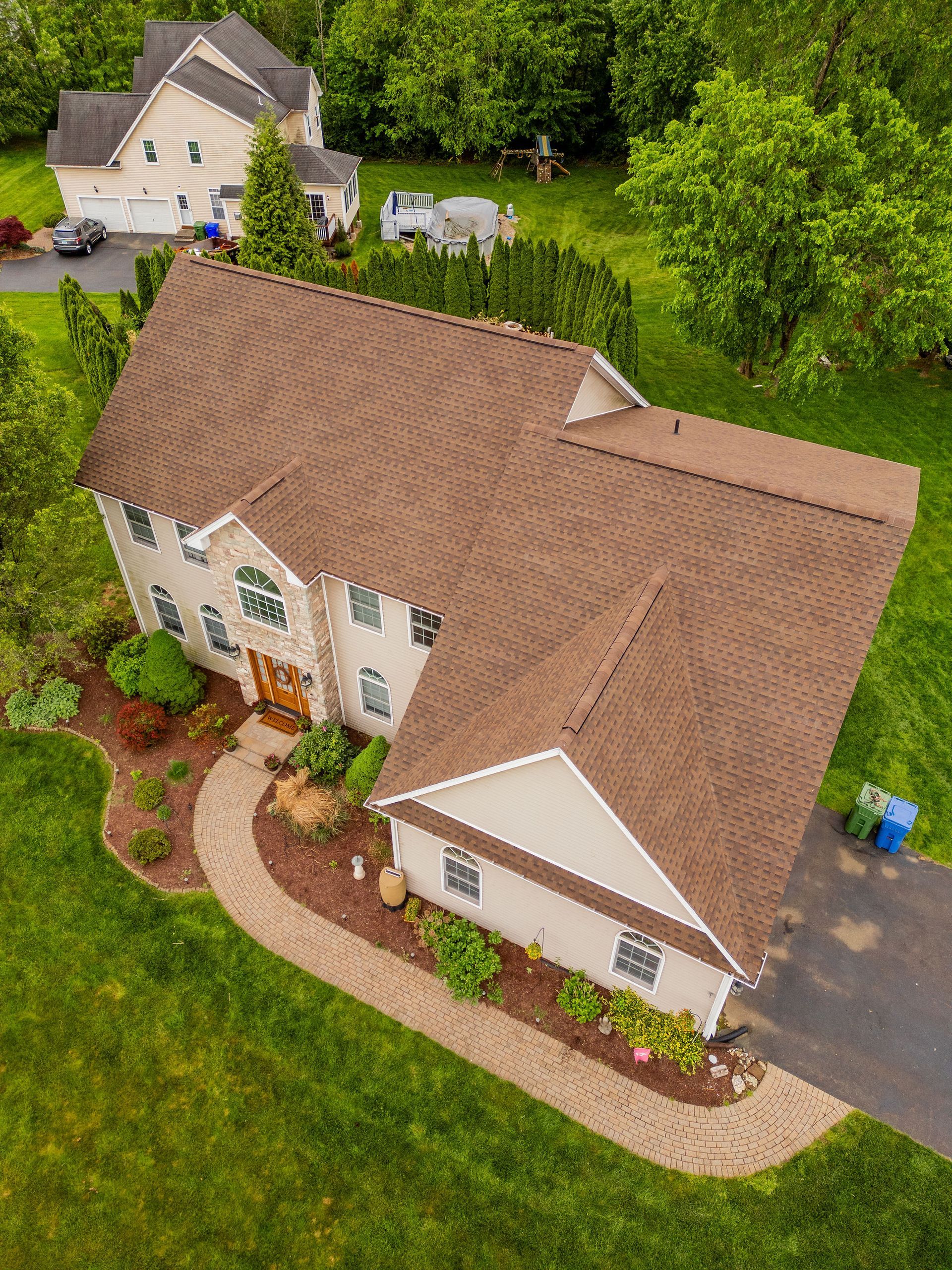 Aerial view of a two-story house with brown roof, beige siding, surrounded by green lawn and trees.