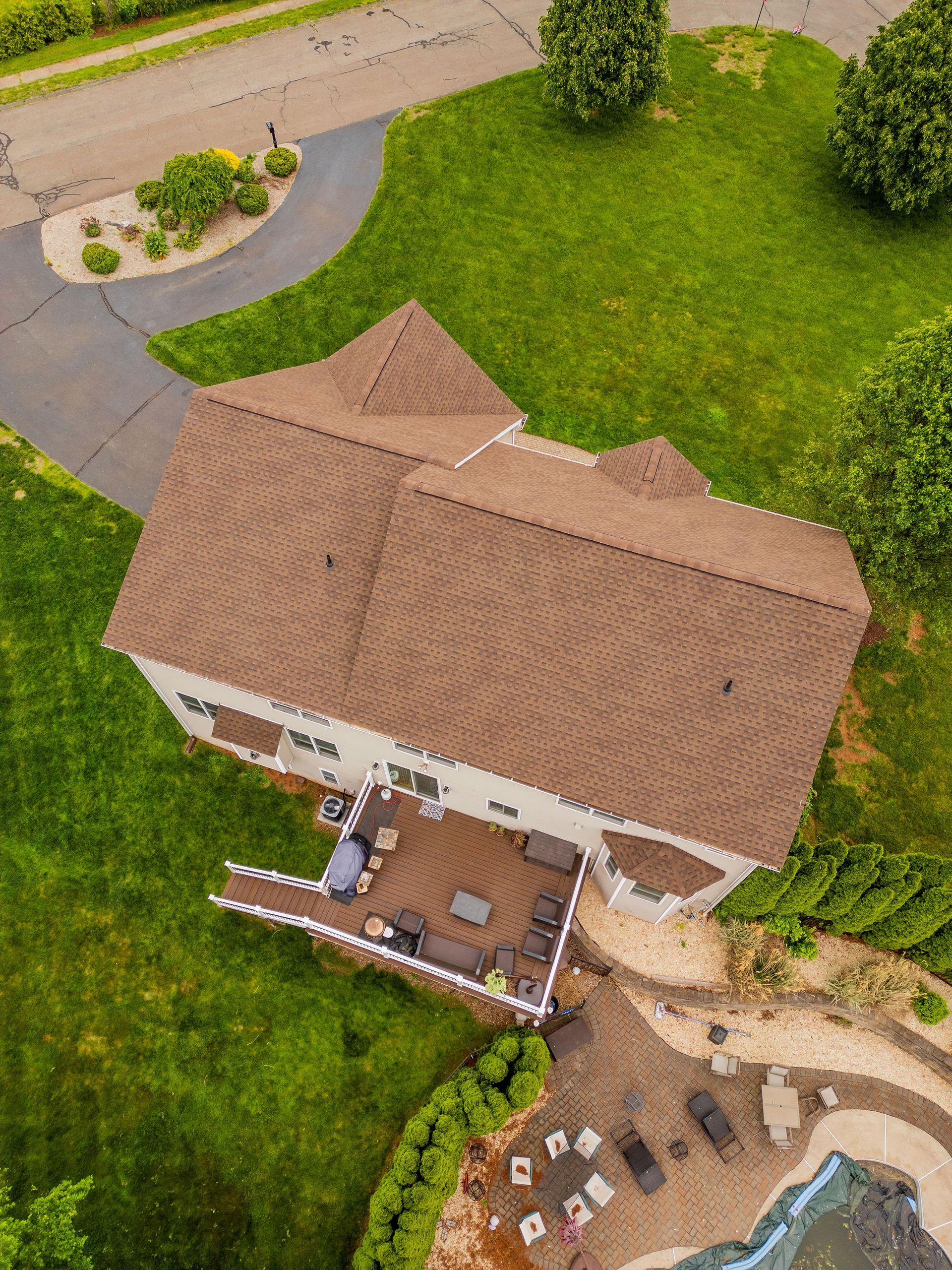 Overhead view of a house with brown roof, deck, and green lawn; driveway and trees in the setting.