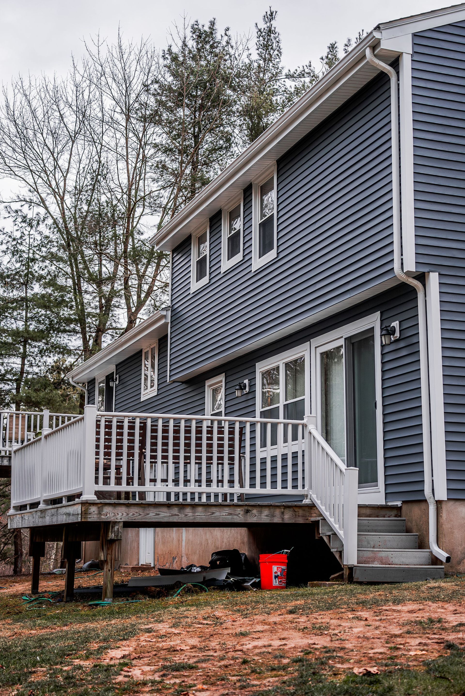 Blue-sided two-story house with white deck and stairs, red bucket at base, trees in background.
