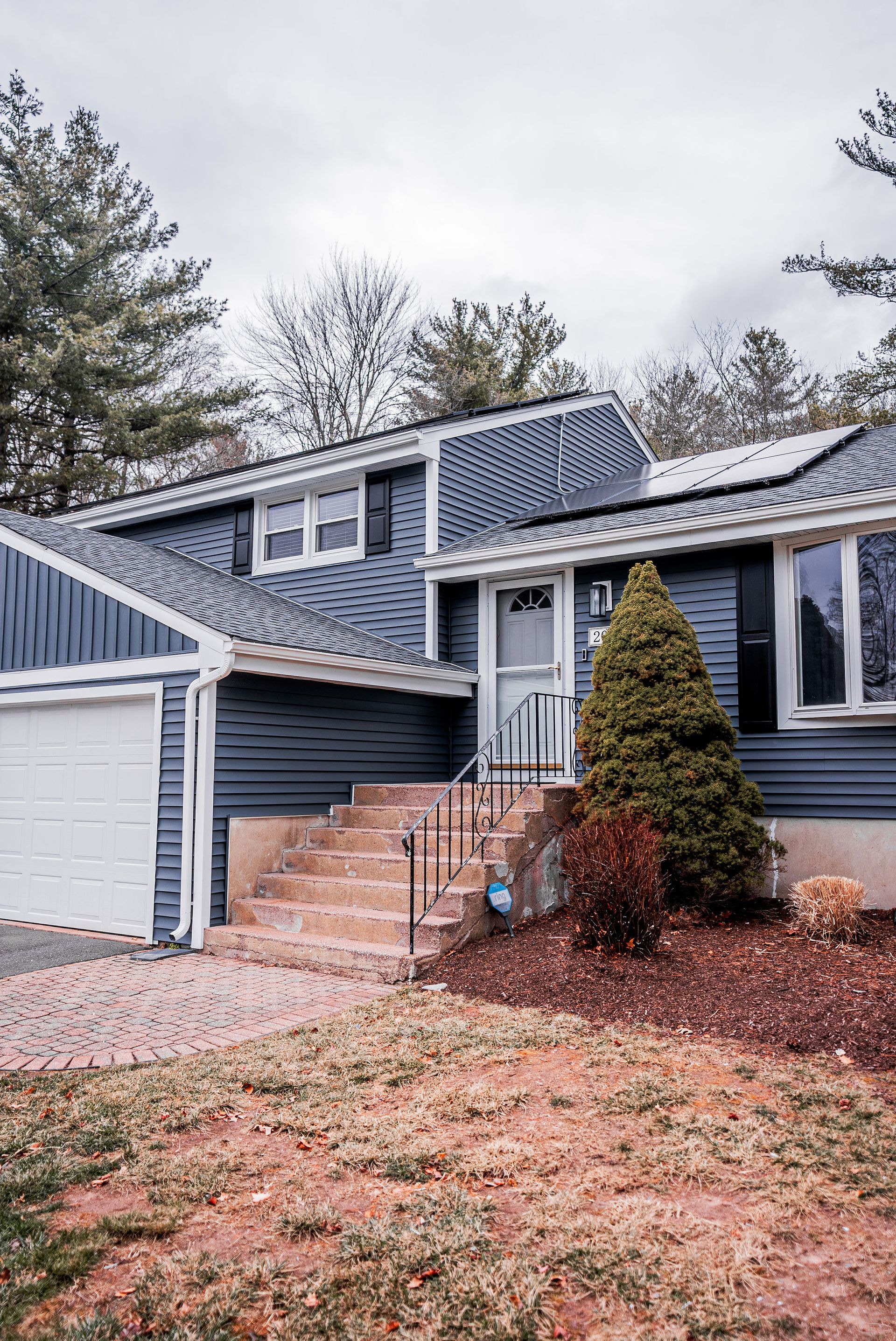 Two-story blue house with white garage, steps to front door, evergreen tree, and brown mulch.