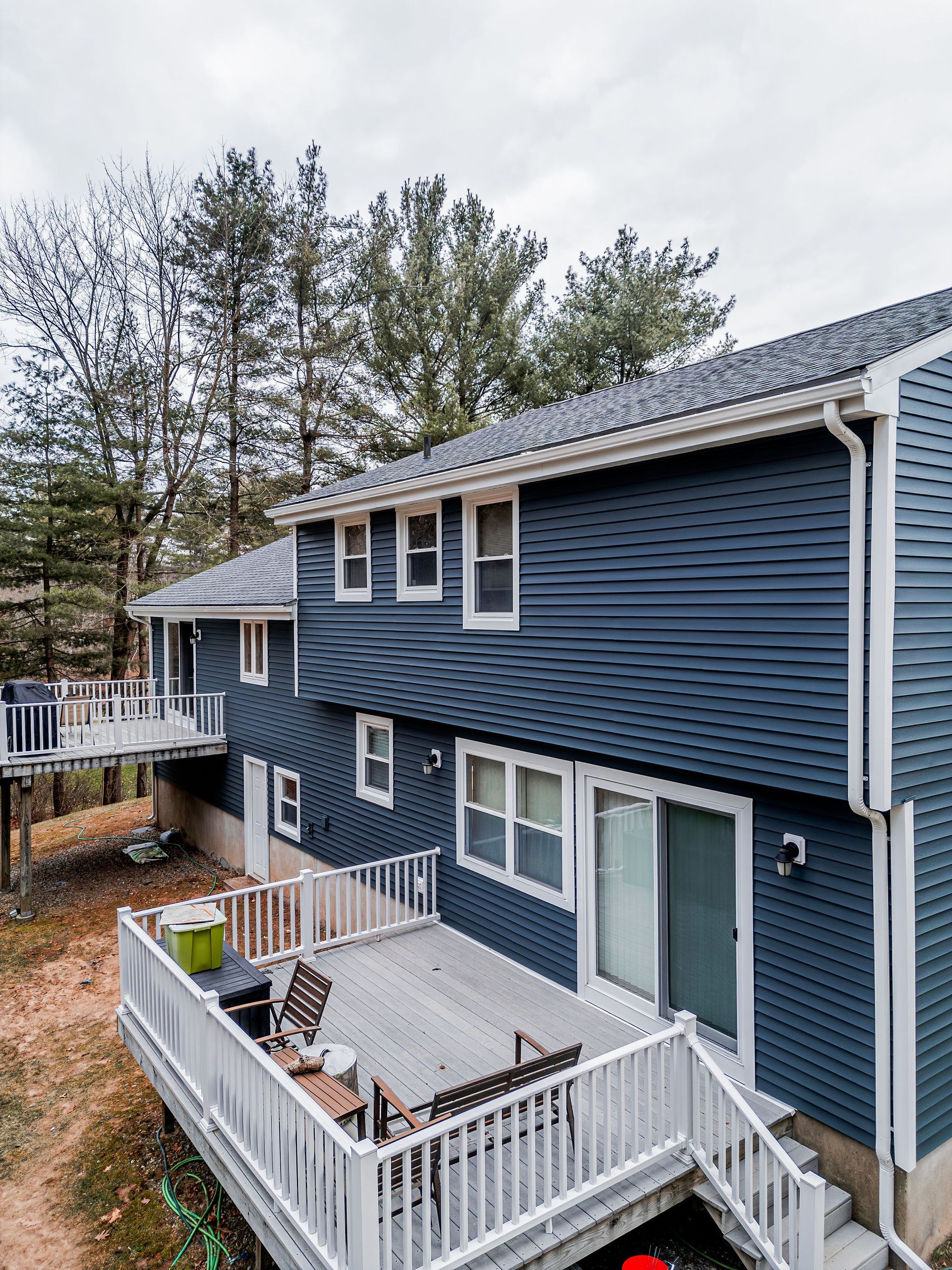 Two-story blue-sided house with decks, white railings, windows, and a sliding glass door; surrounded by trees.