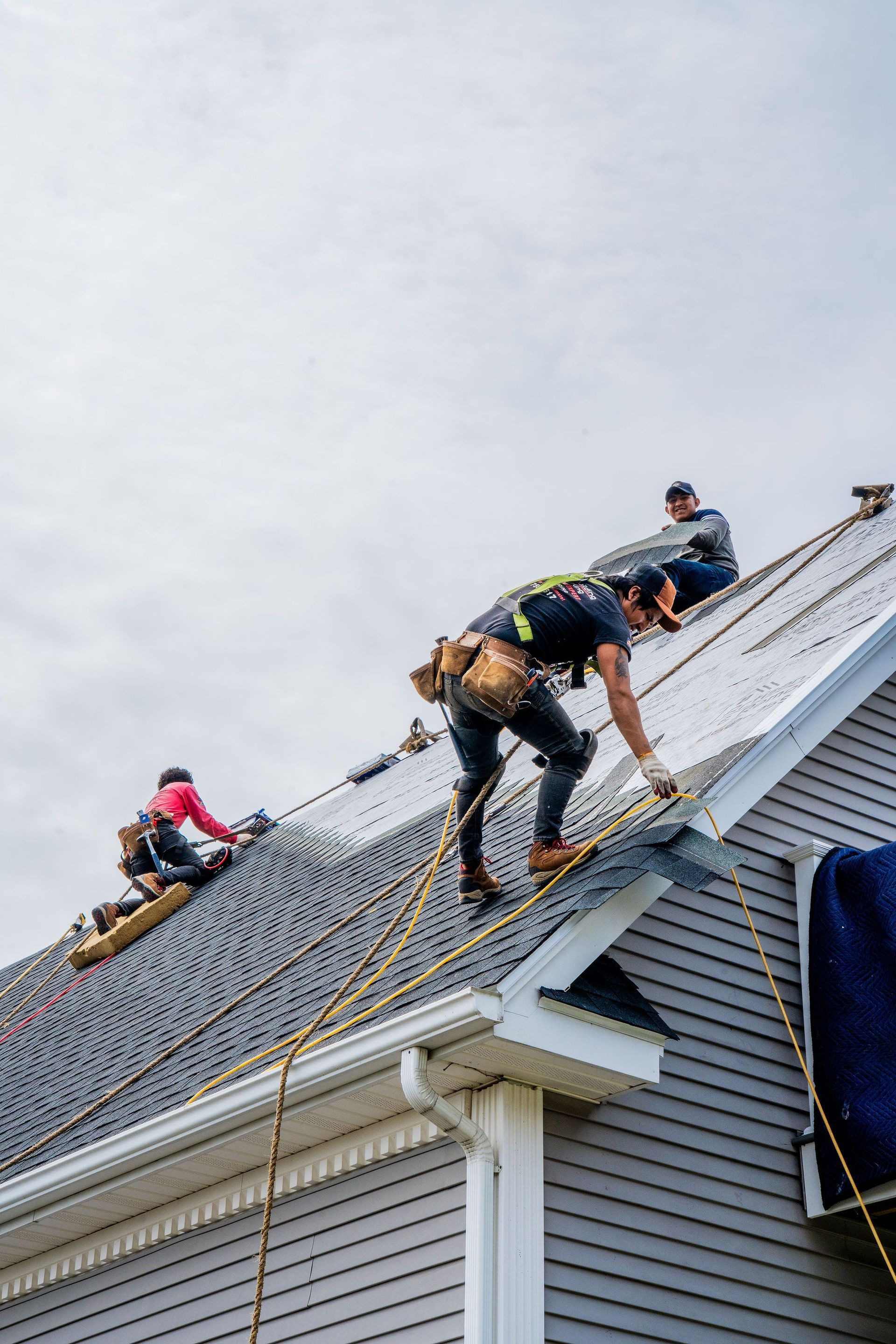 Roofers working on a gray shingled roof, secured with ropes and harnesses. Cloudy sky.