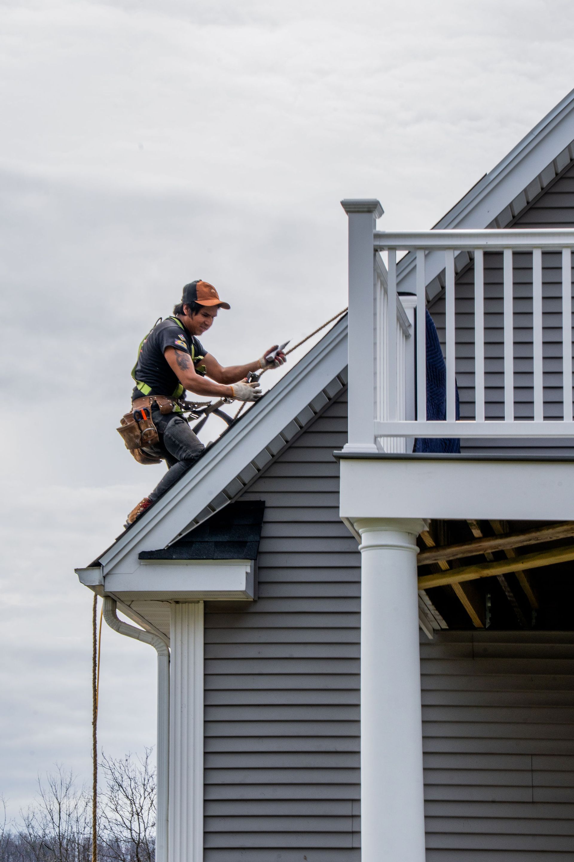A worker on a gray roof, secured by a rope, repairs the eaves of a house with a white railing.