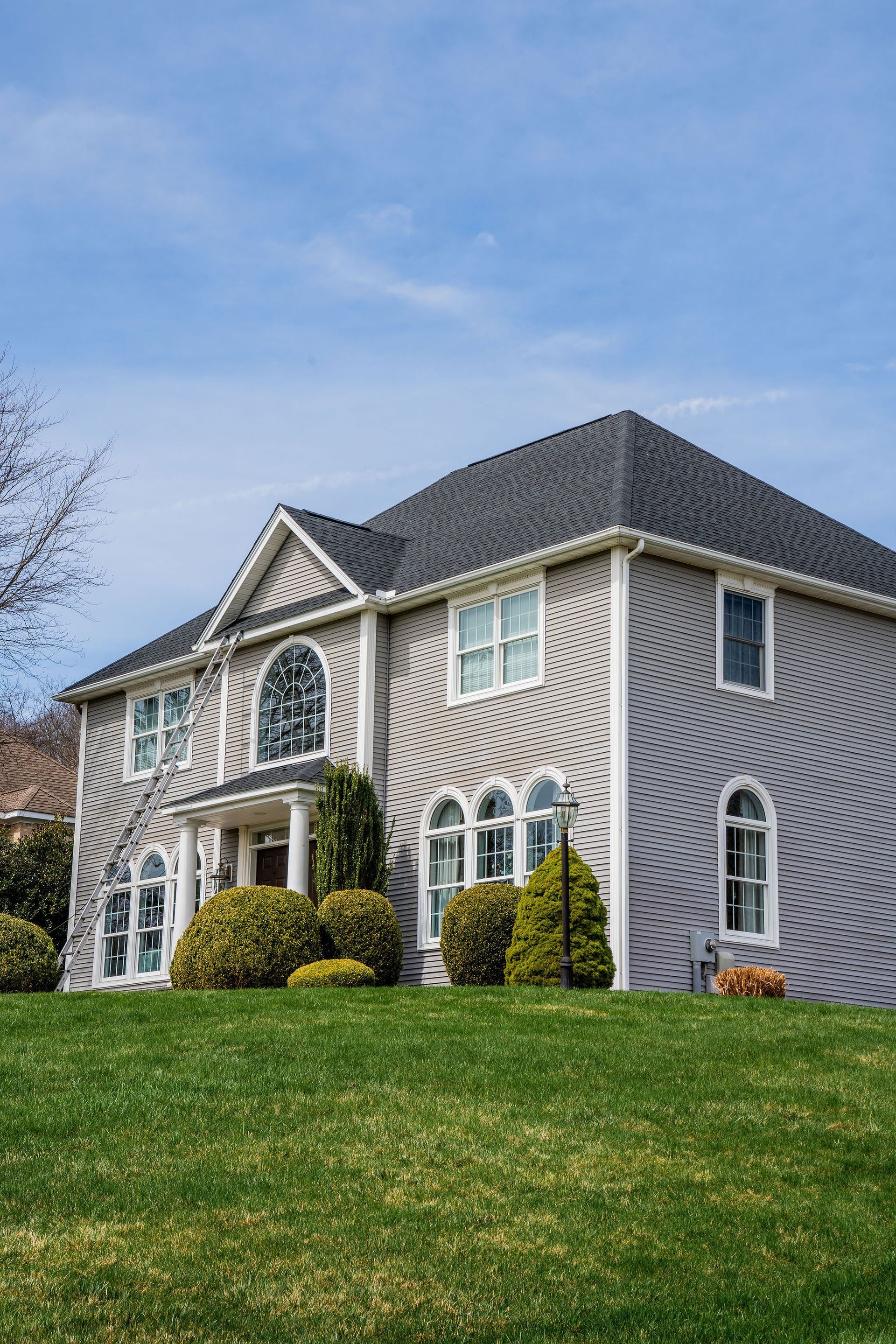 Two-story gray brick house with a dark roof on a grassy hill under a blue sky.