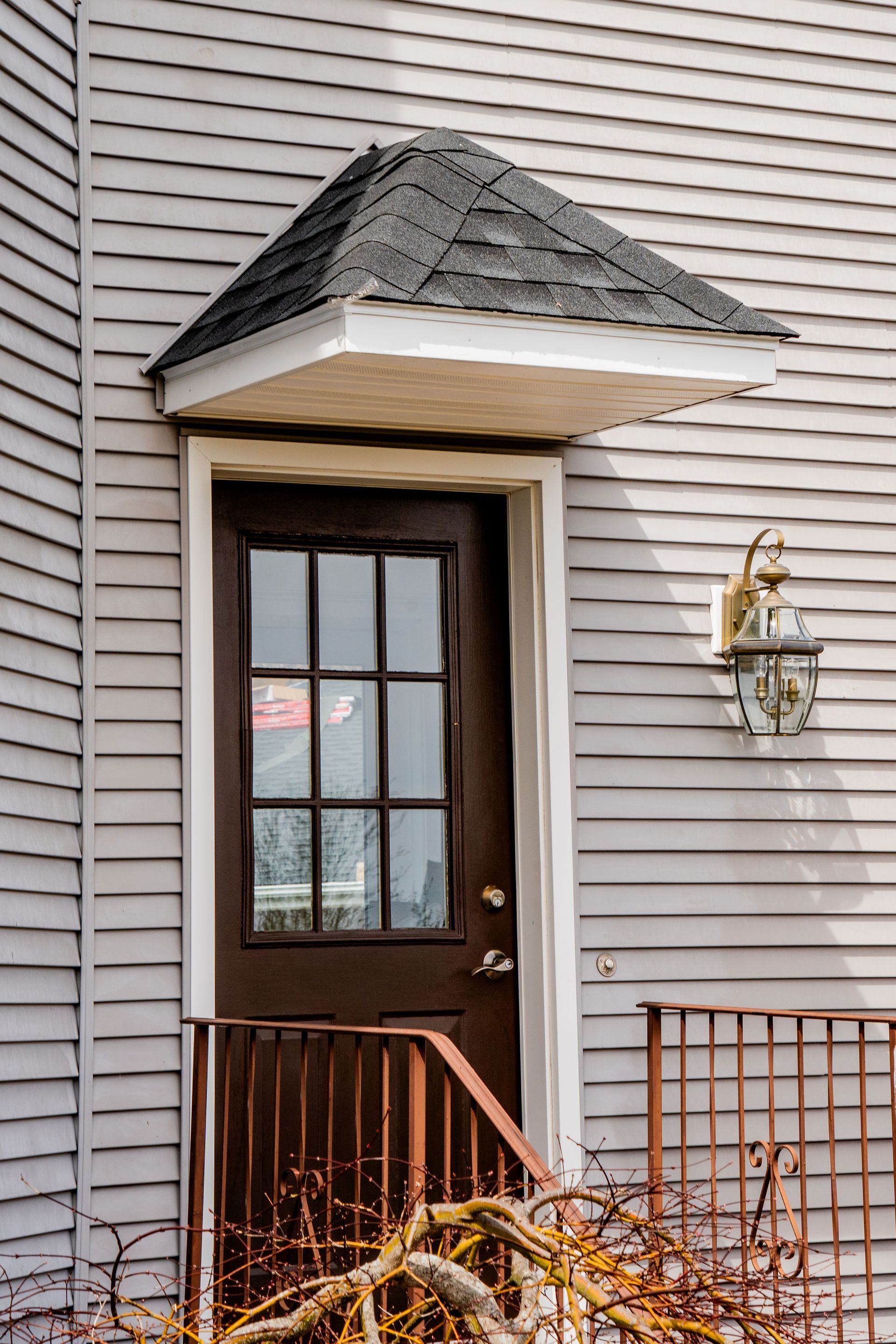 Brown door with multi-pane window under a small roof, flanked by a lamp and railing on a light gray clapboard house.