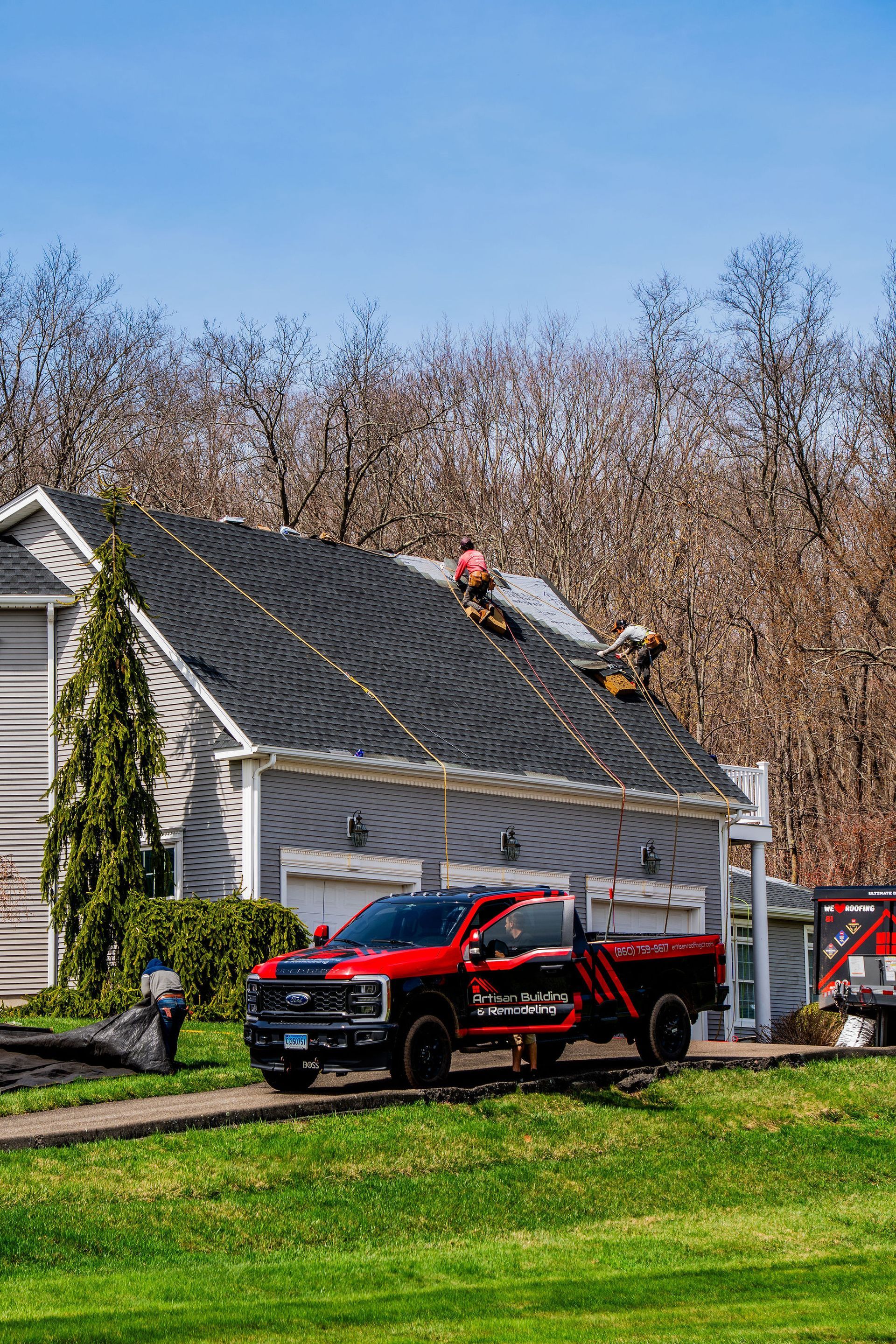 Roofers working on a gray house roof, red truck parked in front. Sunny day.