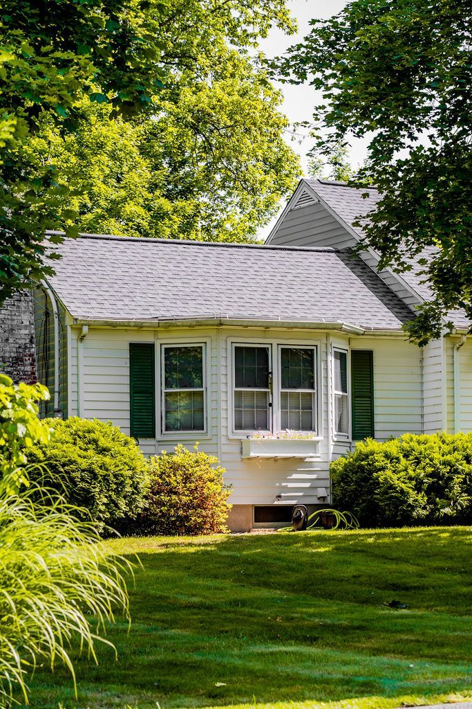 White house with green shutters and a gray roof, surrounded by trees and bushes.