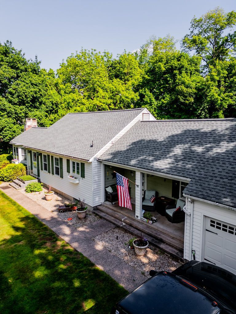 White house with gray roof, American flag hanging on porch, green trees in background.