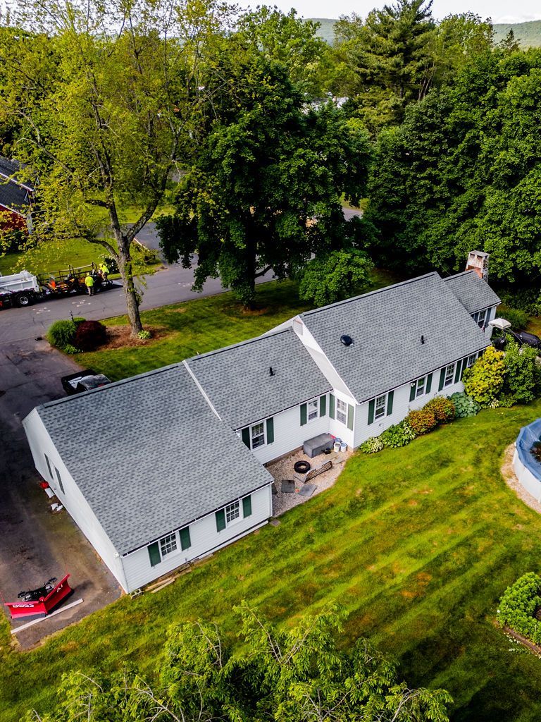 White house with green trim, grey roof, and lush green lawn. A red vehicle and emergency personnel are present.