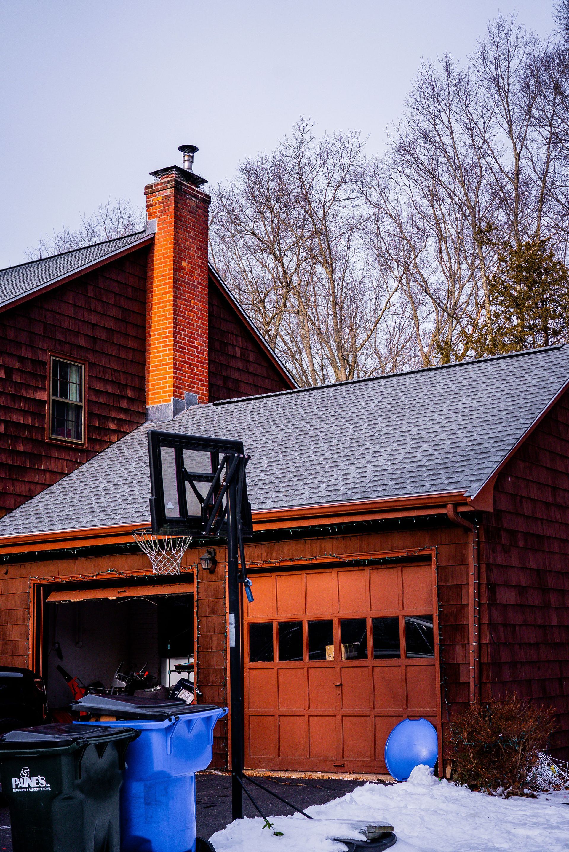 Brown house with open garage, basketball hoop, trash cans, and snow.