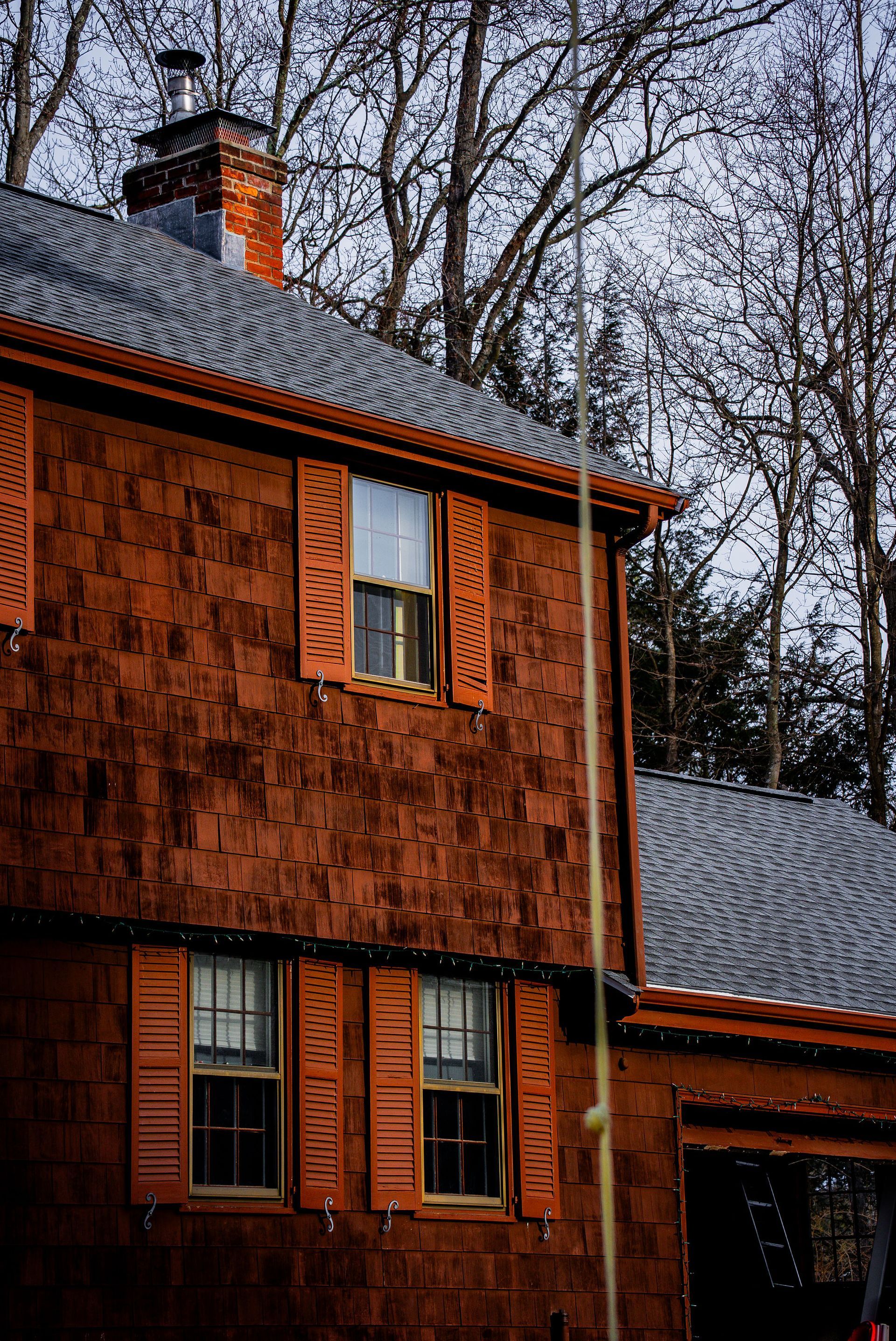 Red-brown shingled house with open shutters, windows, and chimney, set against a cloudy sky and bare trees.