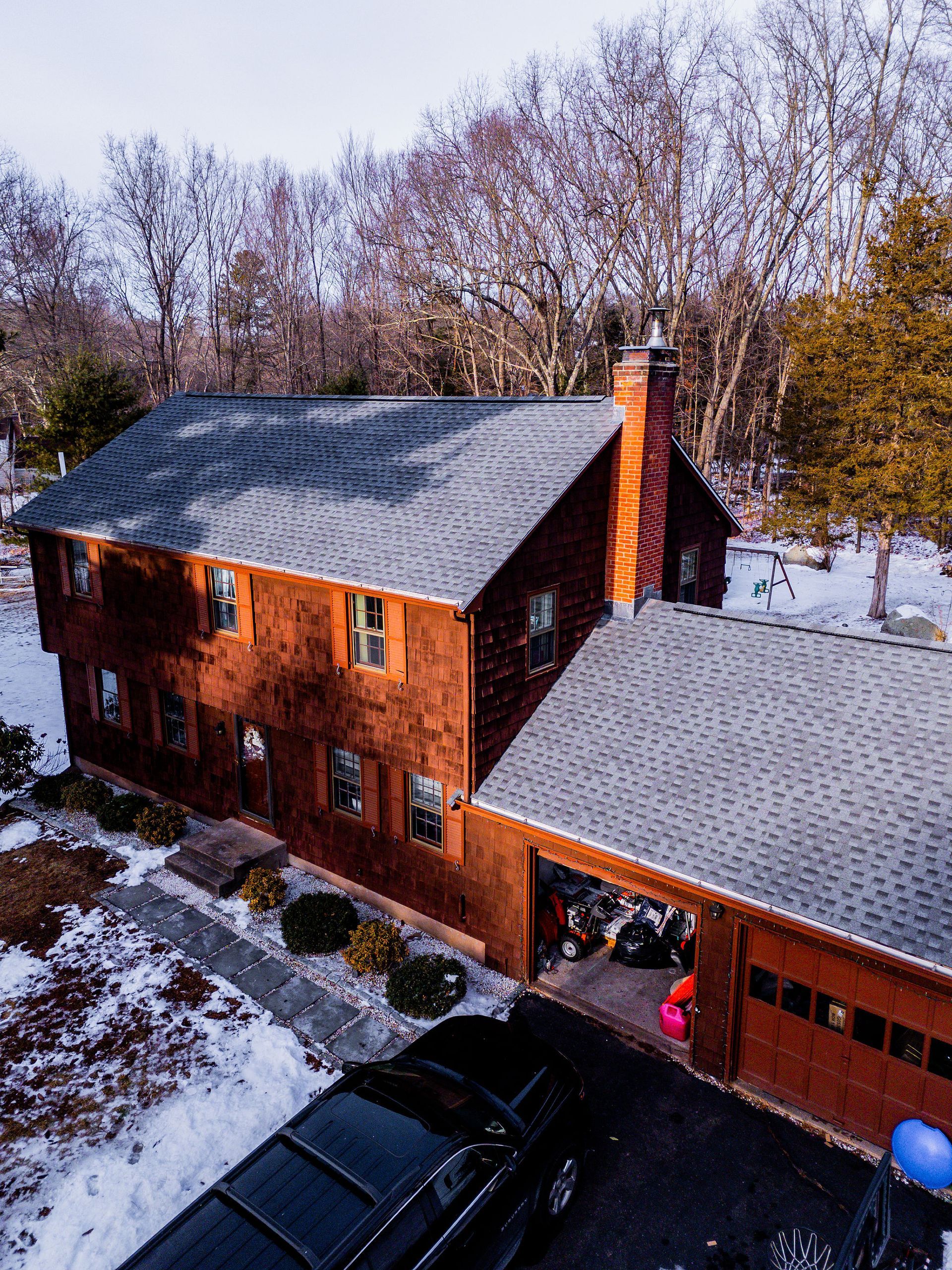 Two-story wood-paneled house with attached garage; snow on the ground; black car in the driveway.
