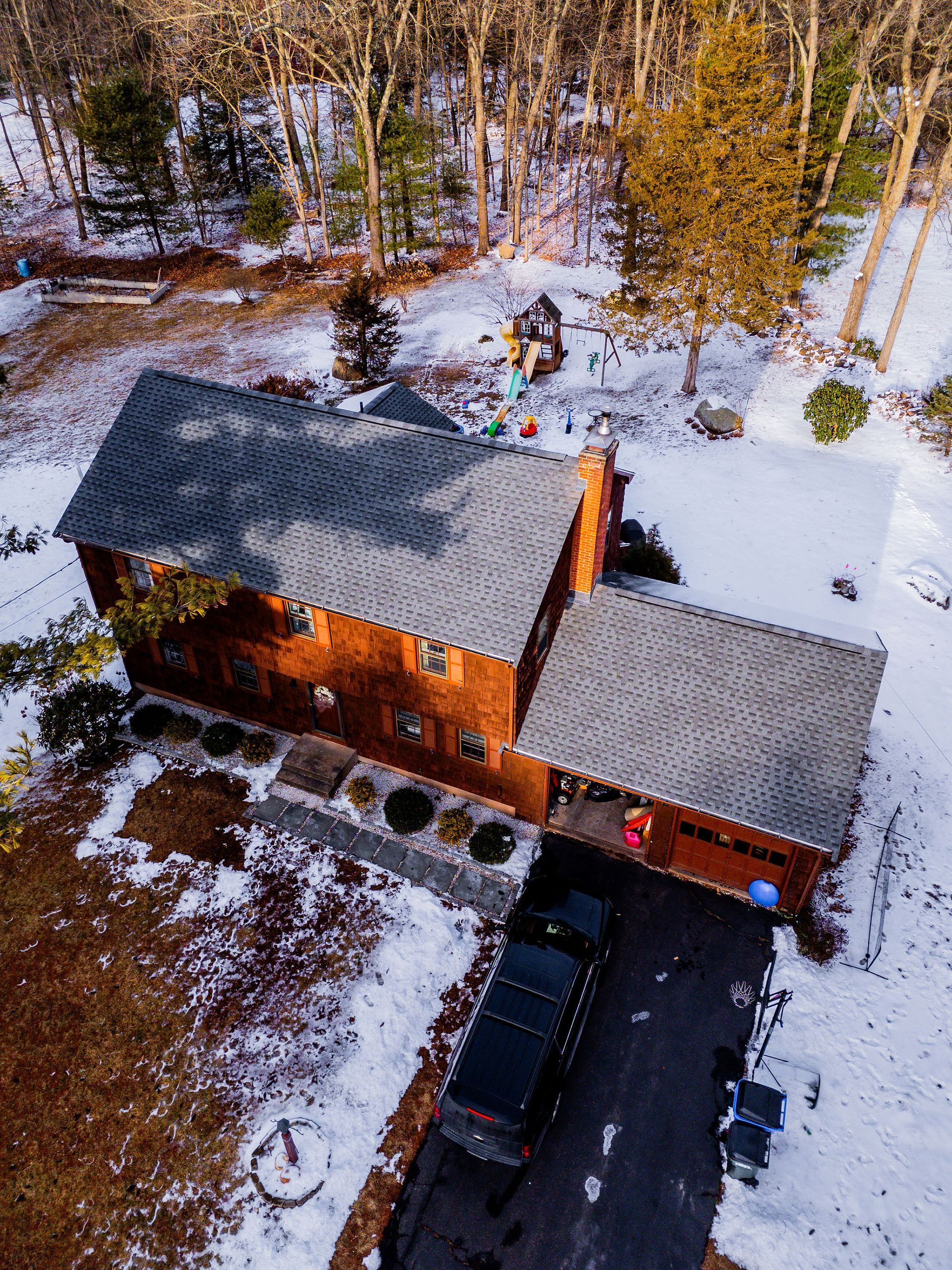 Aerial view of a two-story brick house with a dark driveway in a snowy wooded area.