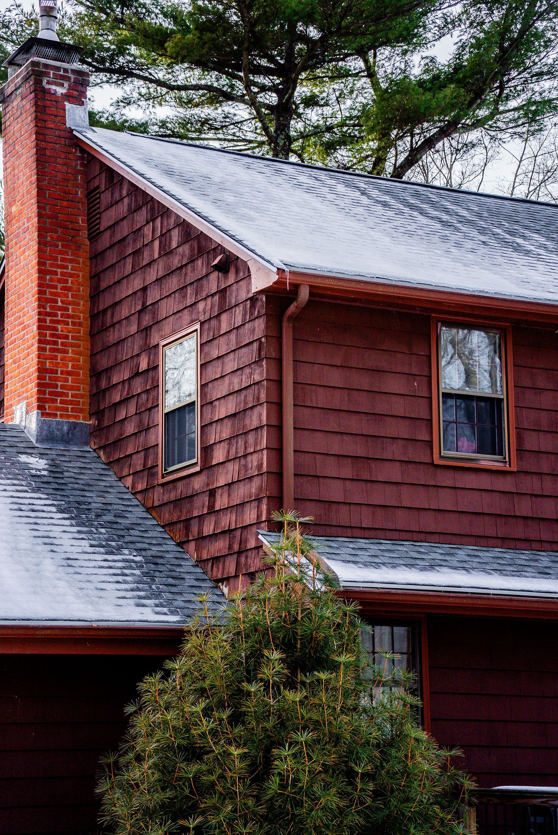 Red-sided house with brick chimney and gray roof, two windows, green bush in front.