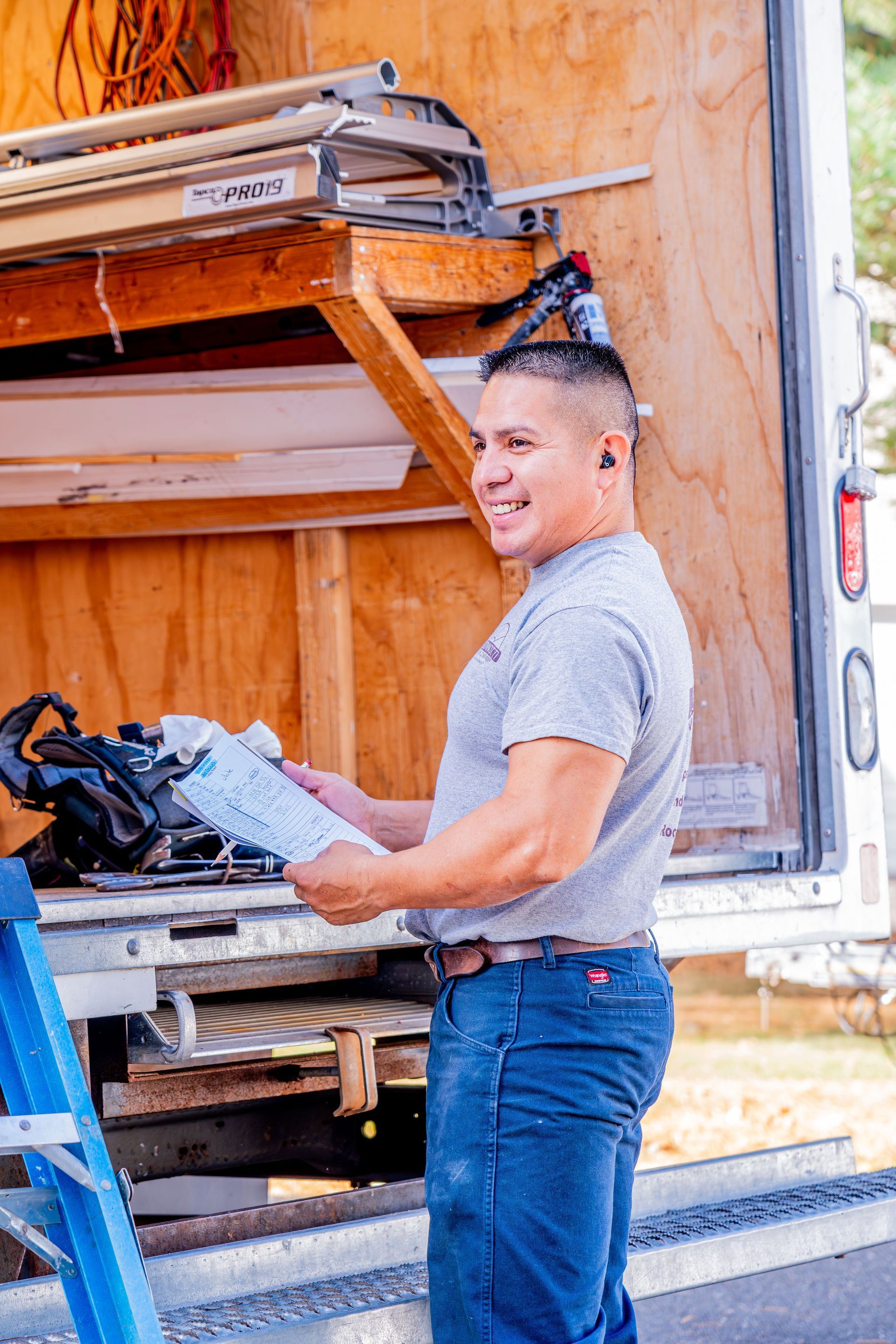 Man standing by a truck holding papers, smiling. He wears a gray shirt, blue jeans, and hearing protection.