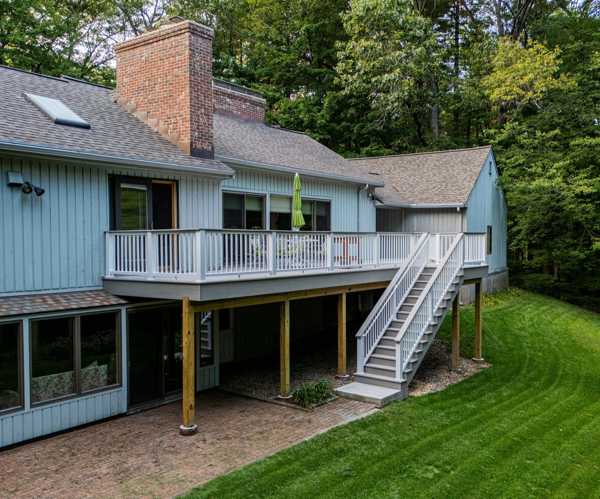 Exterior of a two-story light blue house with a white deck, stairs, and a brick chimney, surrounded by trees.