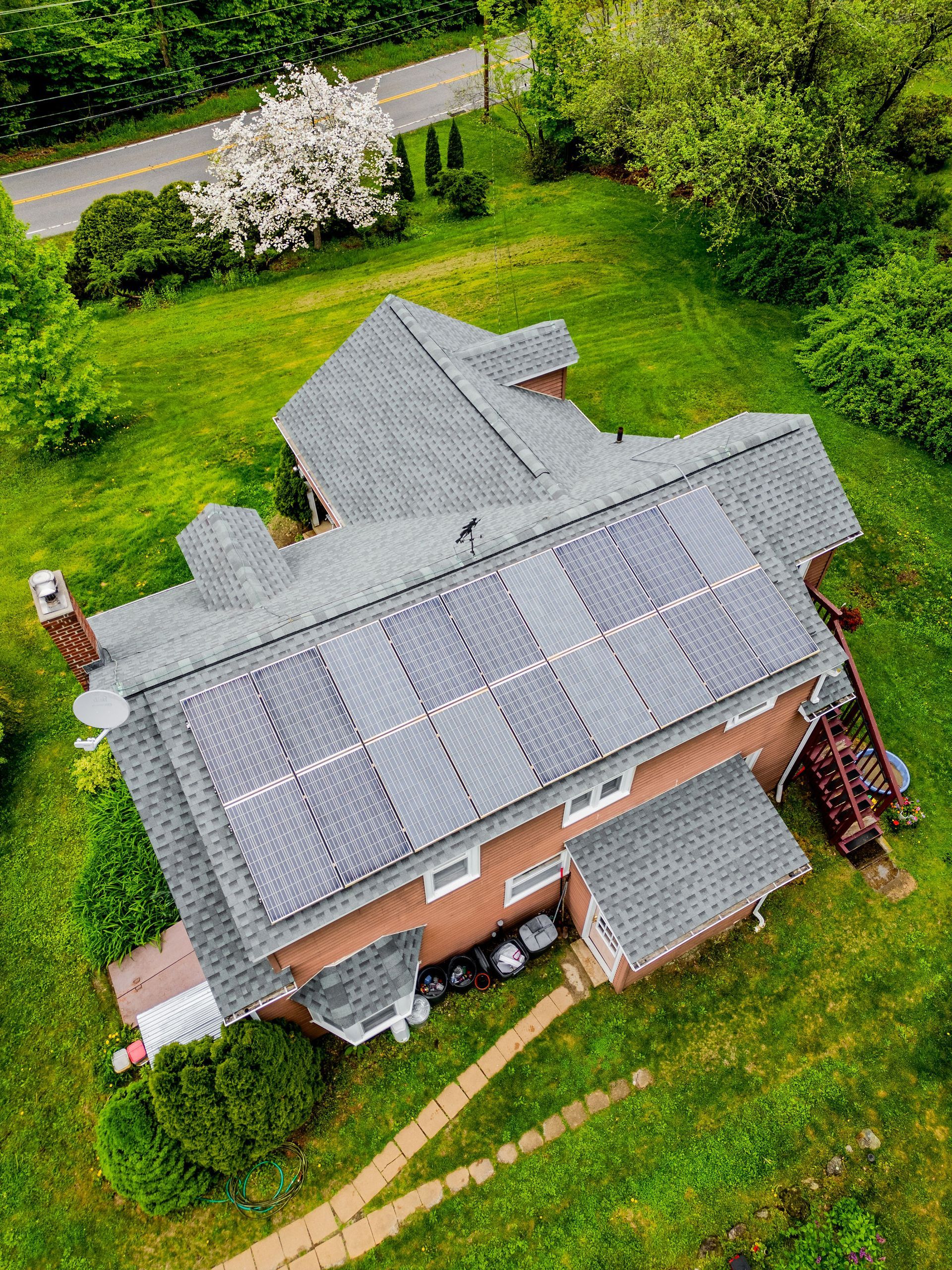 House with solar panels on the roof, surrounded by green grass and trees. A winding road is visible in the background.
