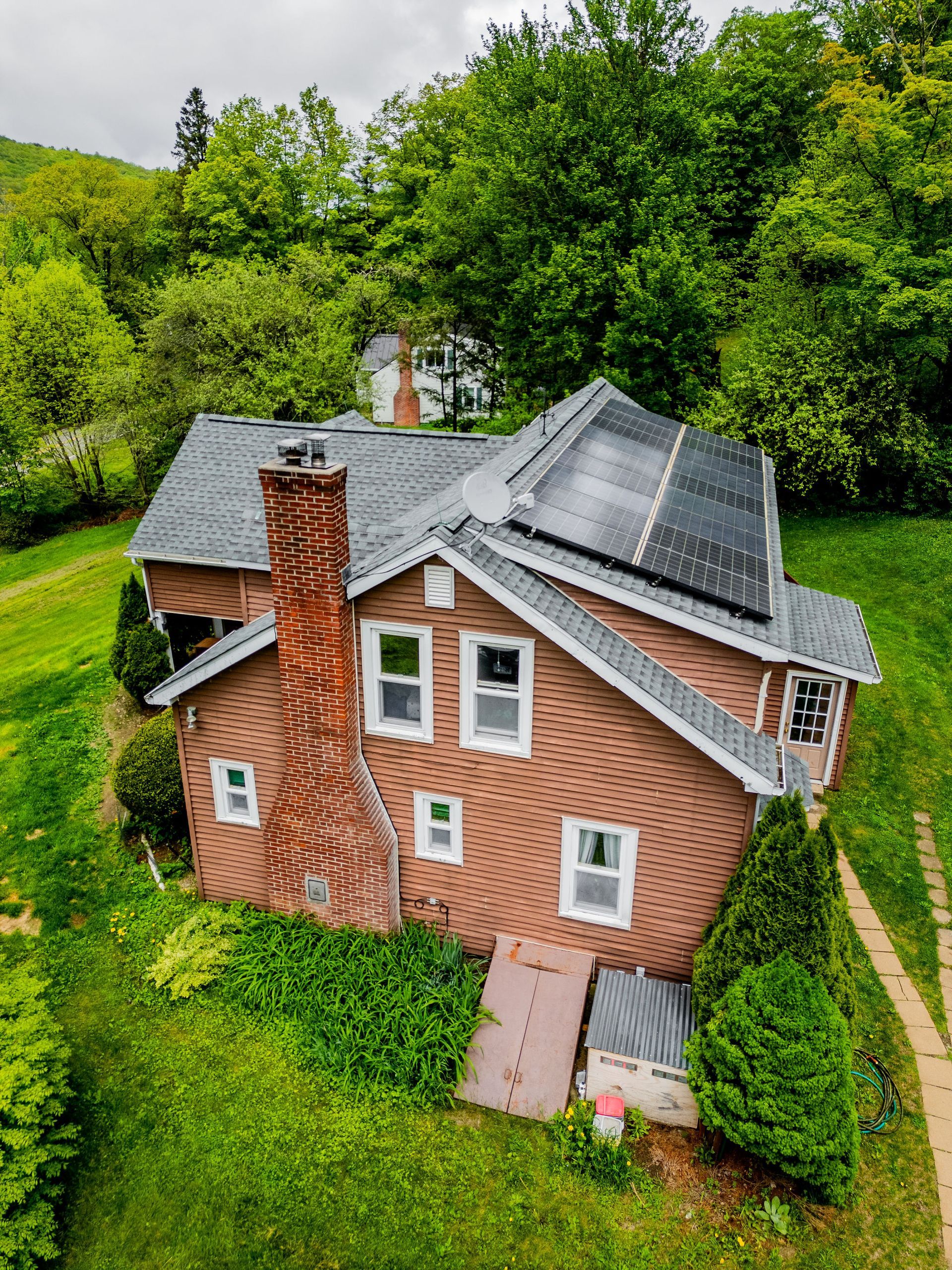 Brown house with solar panels on roof, red brick chimney, set on a grassy hill with trees.