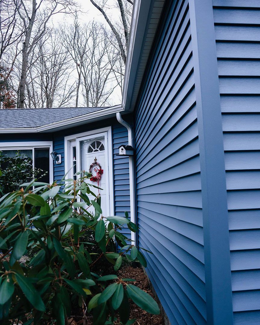 Blue-sided house with white door and wreath. Green bushes in front.