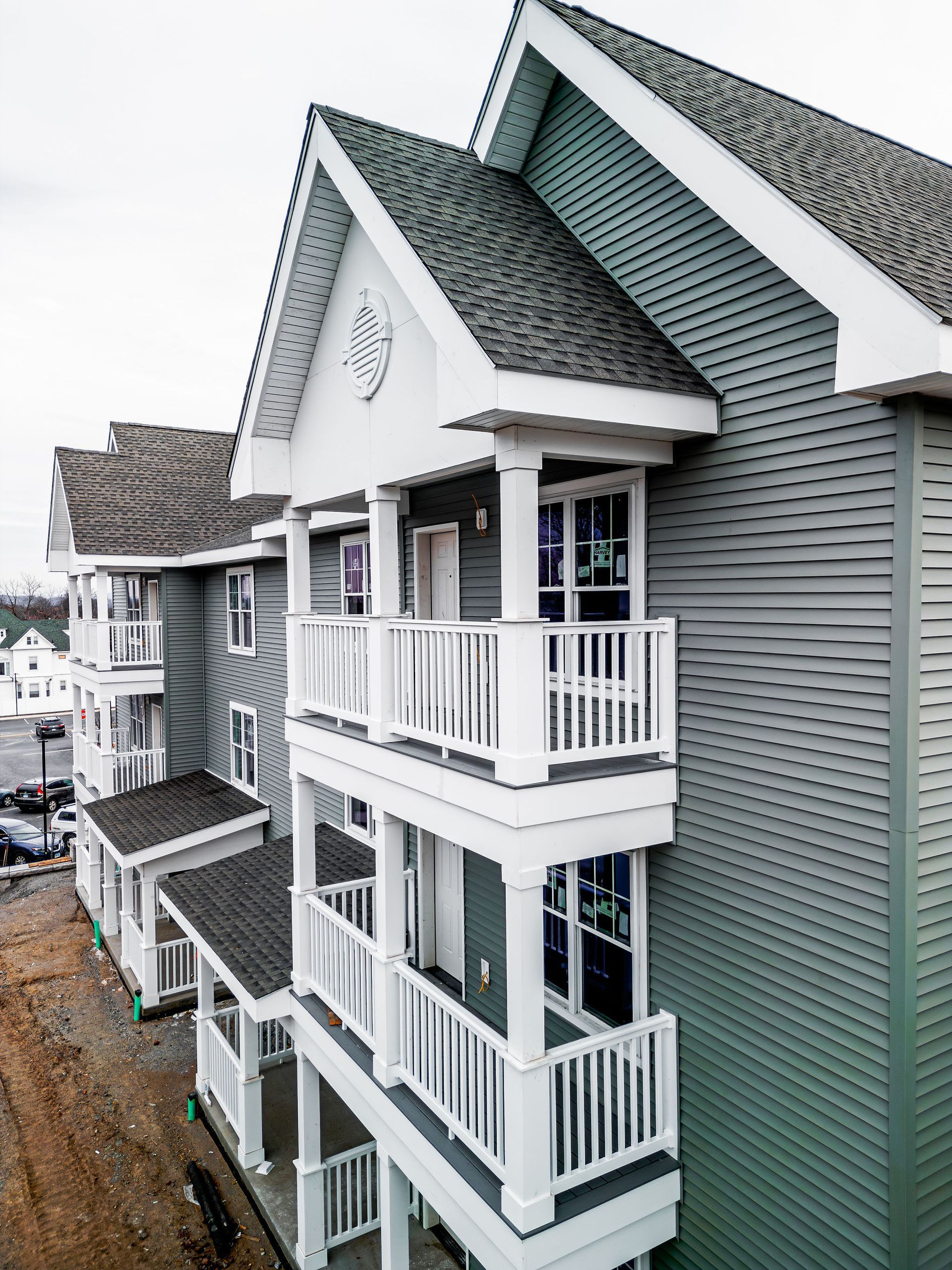 Three-story apartment building with gray siding, white balconies, and a light gray roof under a cloudy sky.