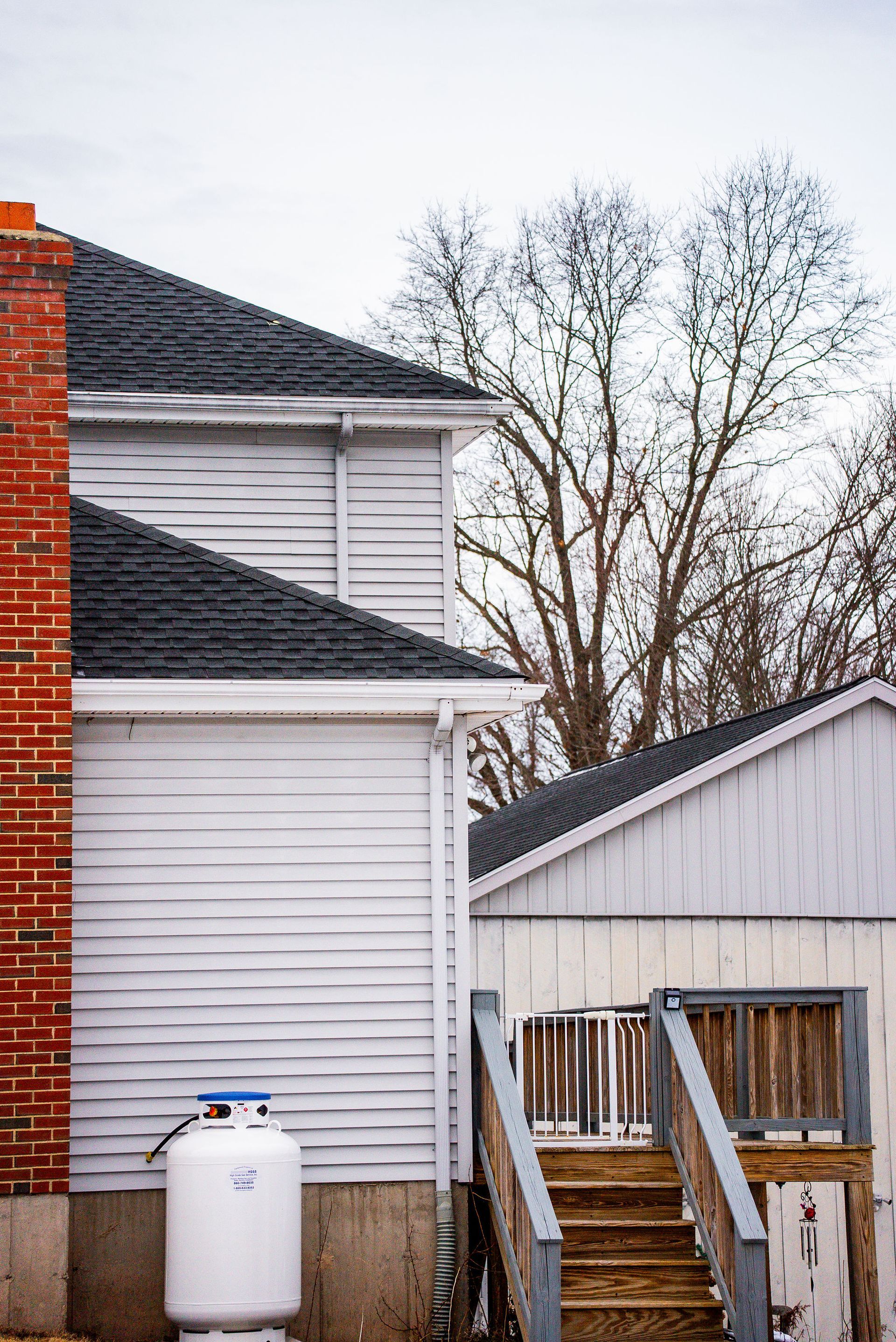 Side of a house with brick chimney, white siding, propane tank, and wooden stairs; leafless trees in background.